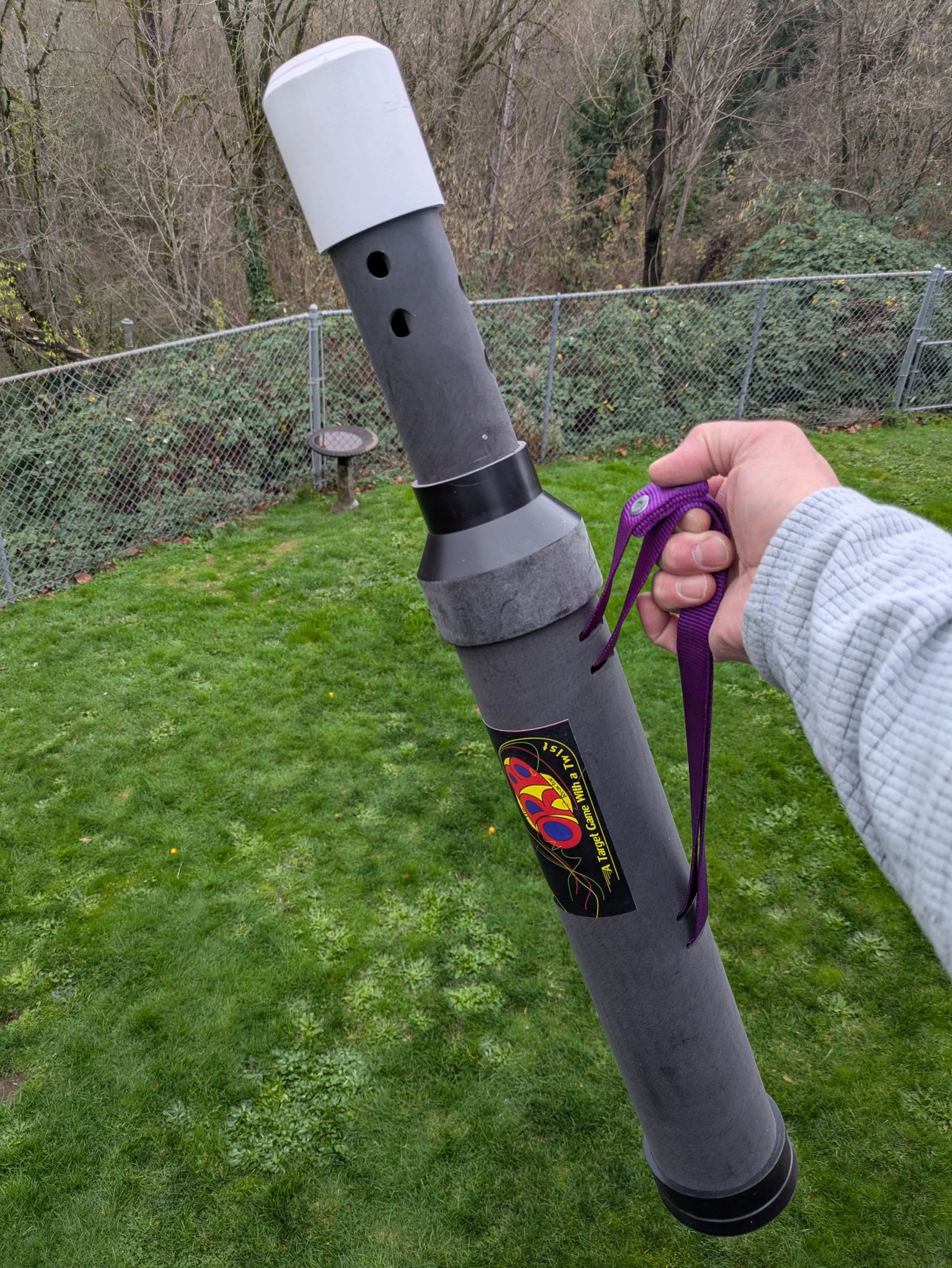 A person holding a rocket-shaped firework with a purple strap attached, outdoors on a grassy area with trees and a chain-link fence in the background.