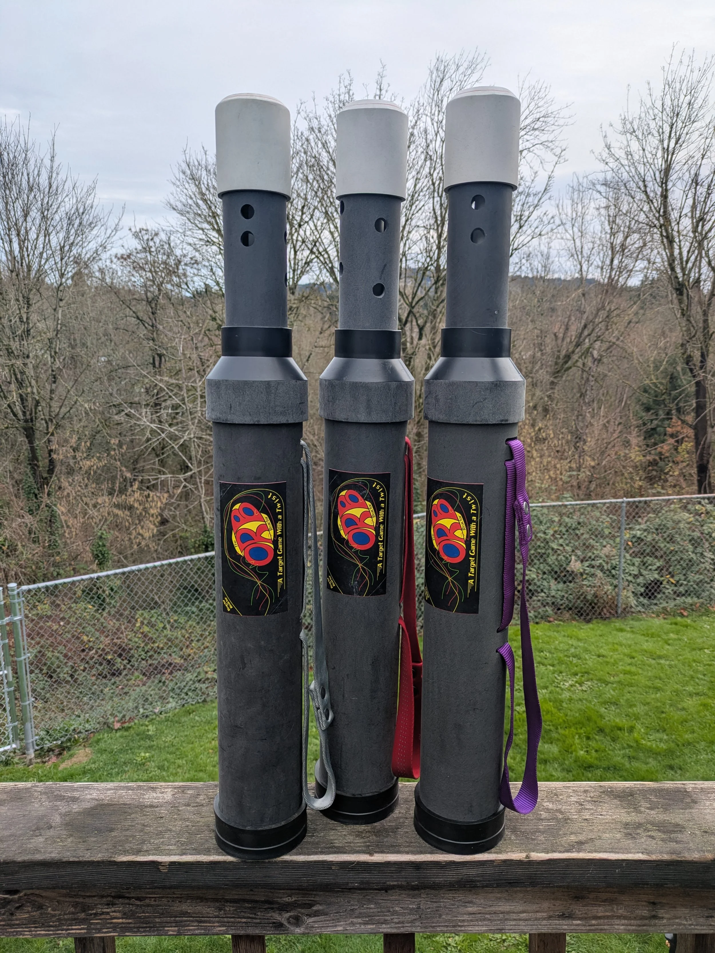 Three vertical cylindrical tubes, with holes and deck flags, stand on a wooden railing outdoors, with trees and a chain-link fence in the background.