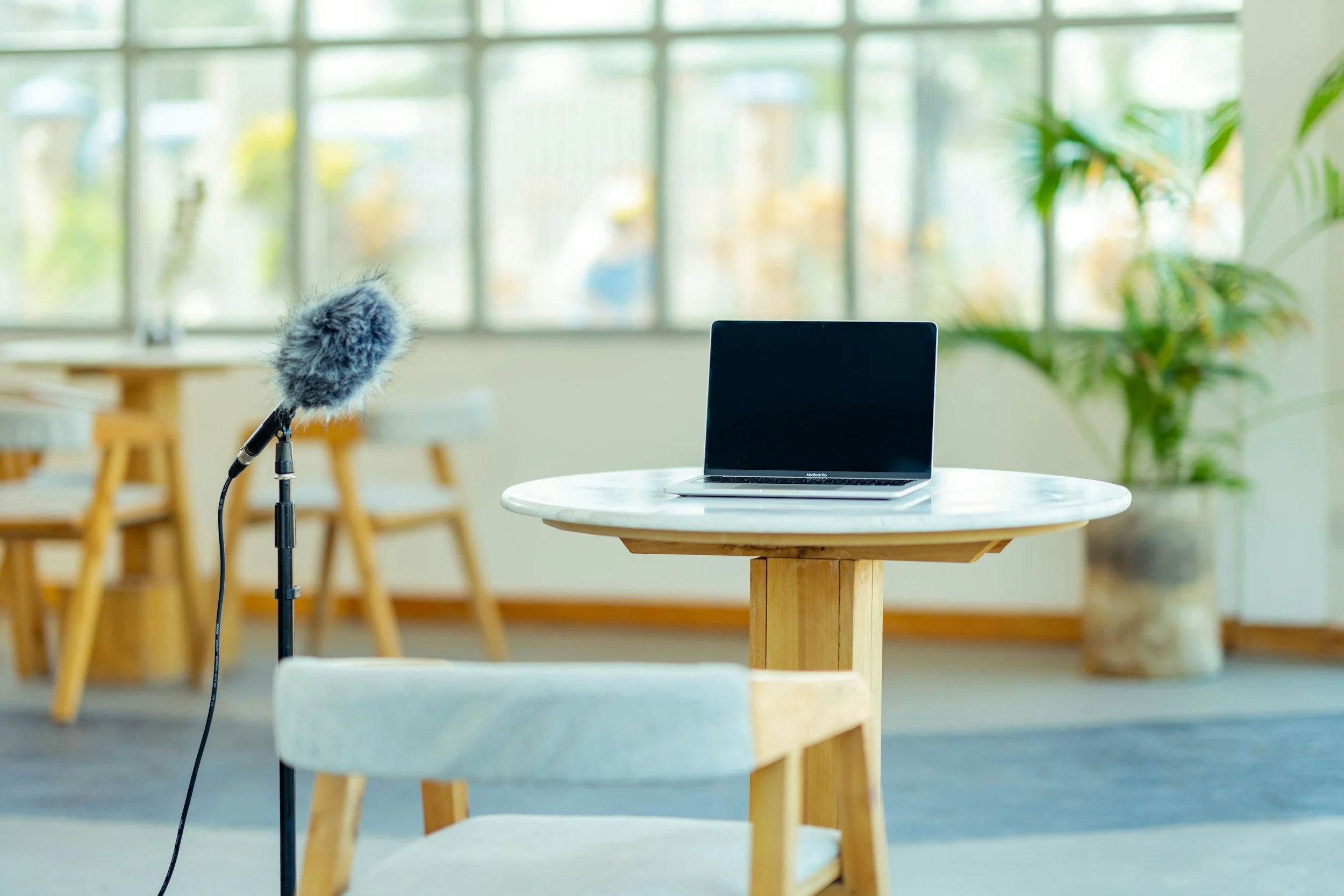 A laptop on a round wooden table with a beige chair in front, a microphone on a stand nearby, in a bright room with large windows and green plants.