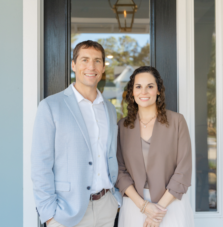 A man and woman standing side by side at the doorway of a house, smiling at the camera.