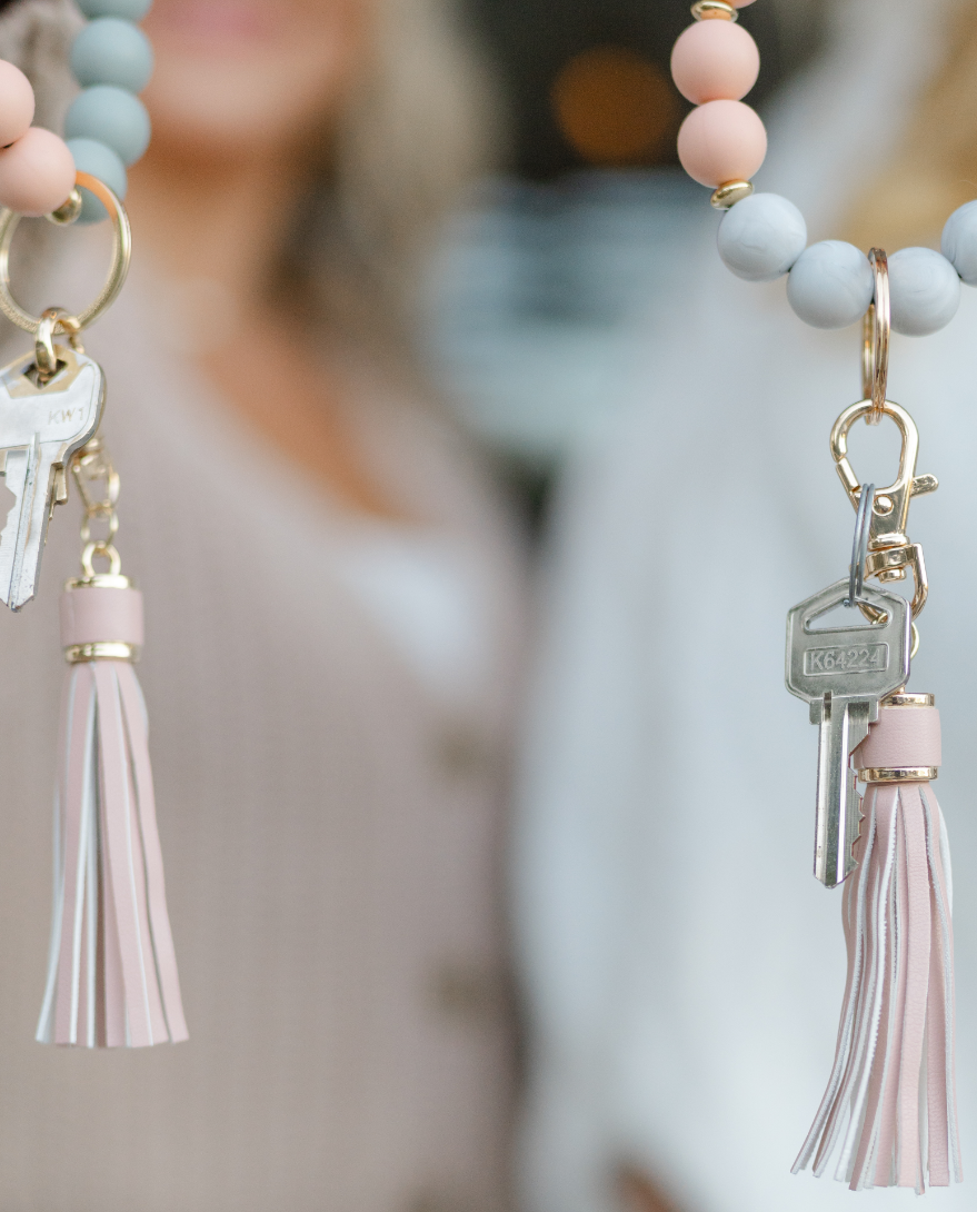 Close-up of pastel-colored beaded lanyards with keys, tassels, and gold accents, held by a person in a beige and white outfit.