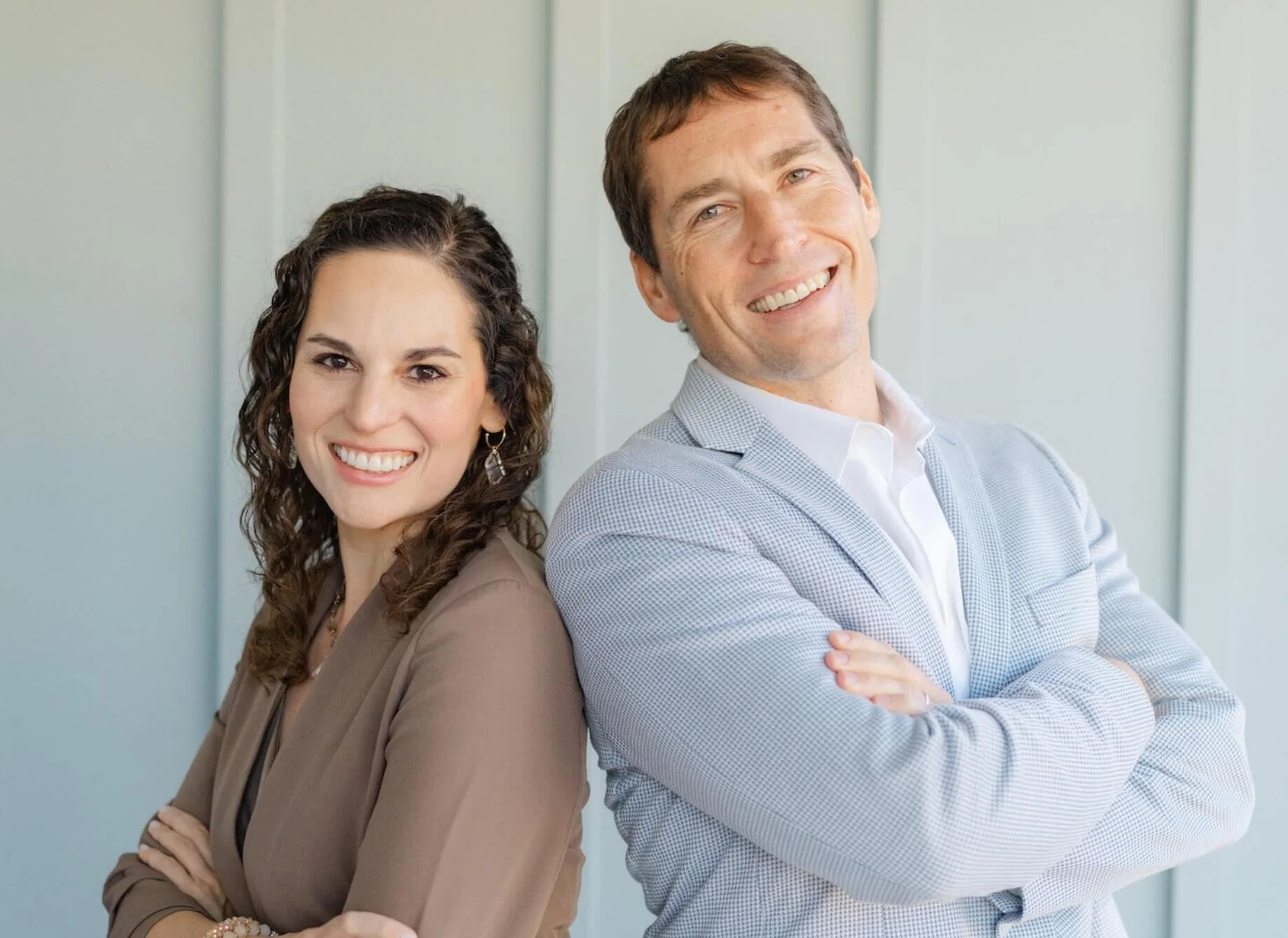 A woman and a man standing back-to-back with arms crossed, smiling at the camera, in front of a light-colored wall.