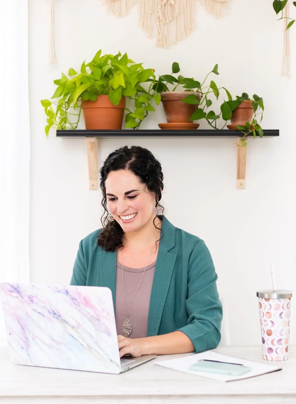 A woman sitting at a desk, smiling at her laptop, with a paper cup and smartphone nearby, in a room with a wall shelf holding three potted plants.