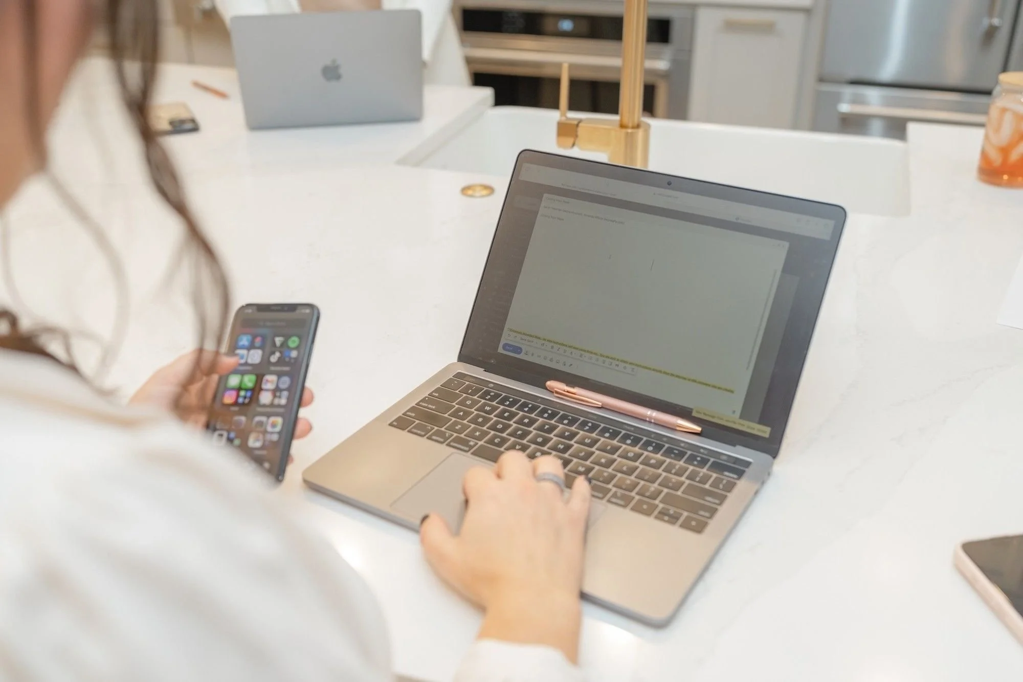 Sarah Newman using a laptop and smartphone at a white kitchen island.