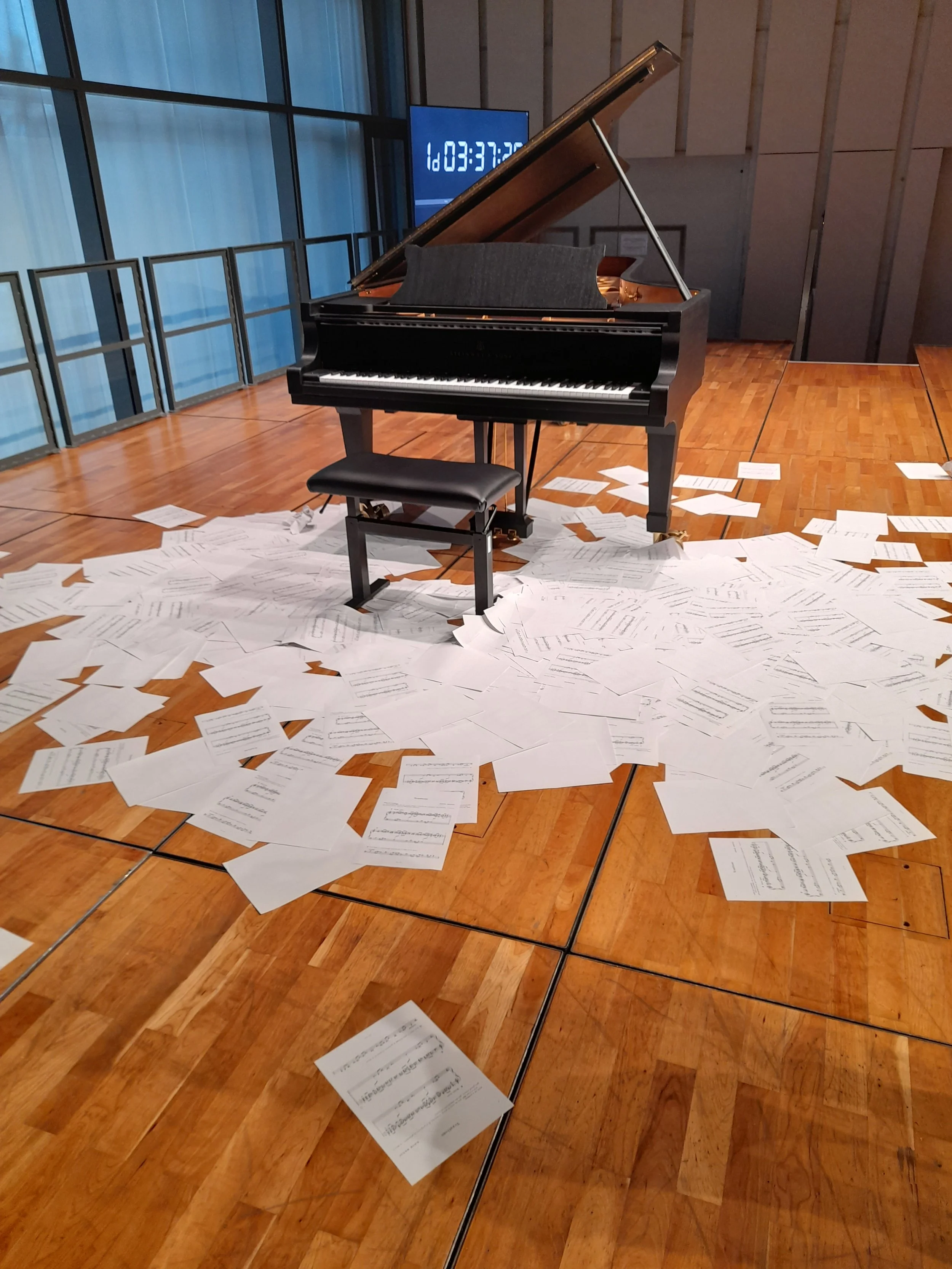 A grand piano on a wooden floor surrounded by scattered sheets of sheet music, with a digital clock showing the time as 03:37 in the background.