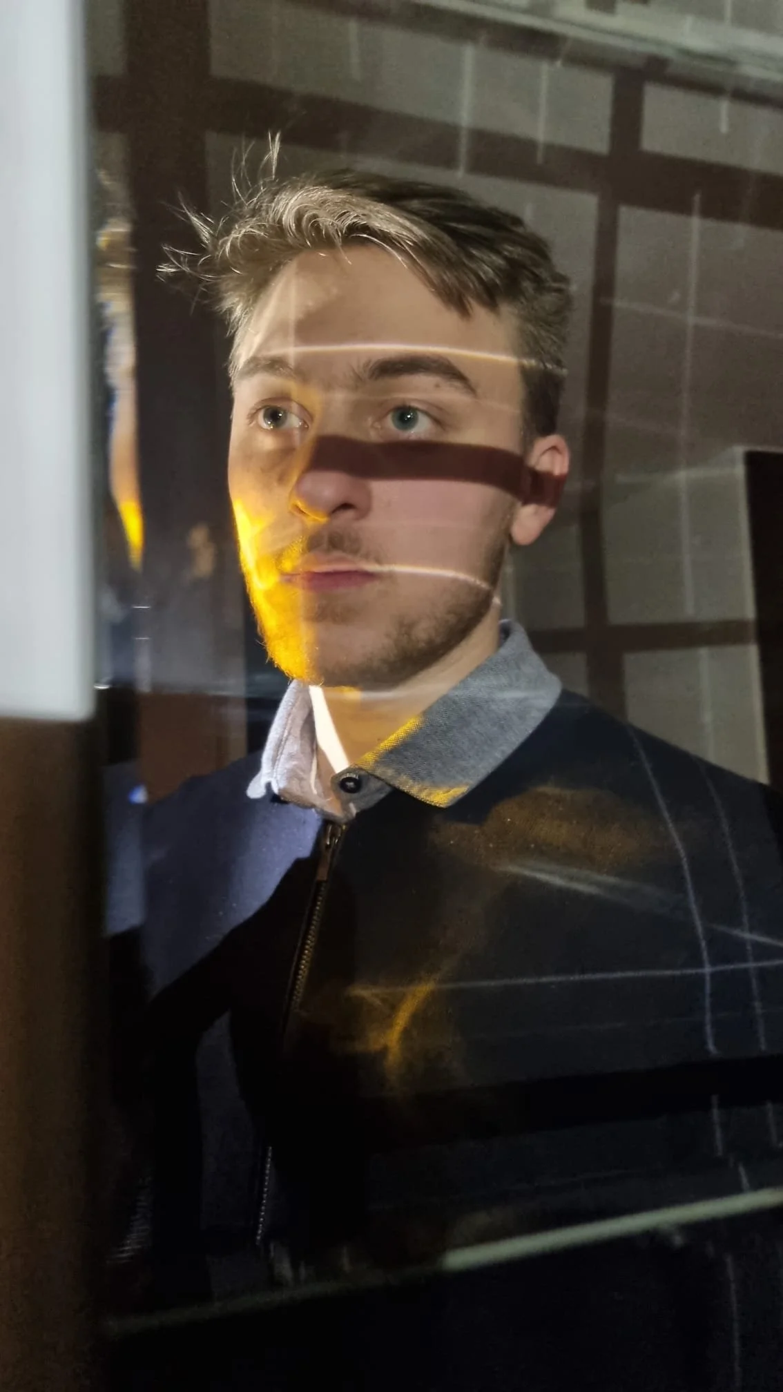 Hugo Daouk in Beirut in 2023. A young man with dark blond hair and blue eyes looking through a glass surface with horizontal light reflections, illuminated by yellow light on one side.