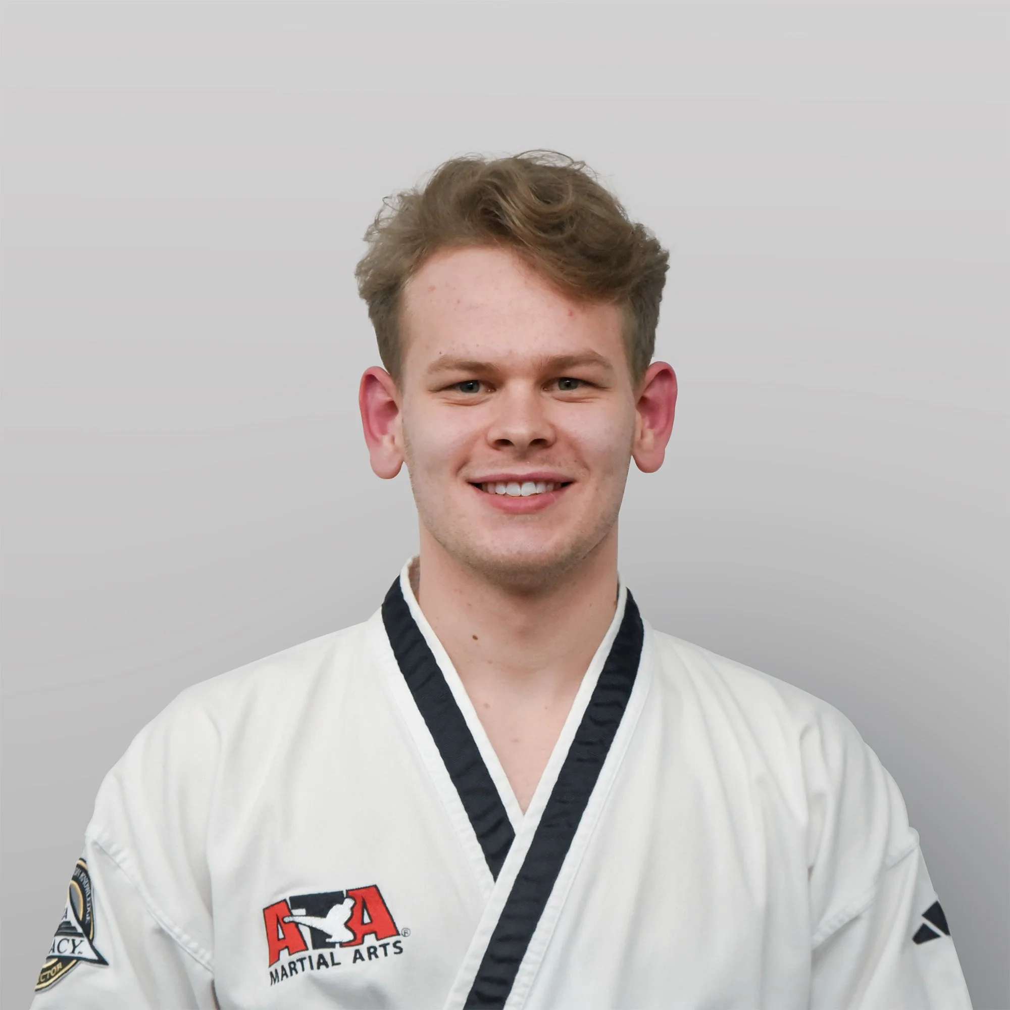 A young man with short, light brown hair, light skin, and a slight smile, wearing a white martial arts uniform with black trim, patches on the sleeves, and an American Taekwondo Association logo.