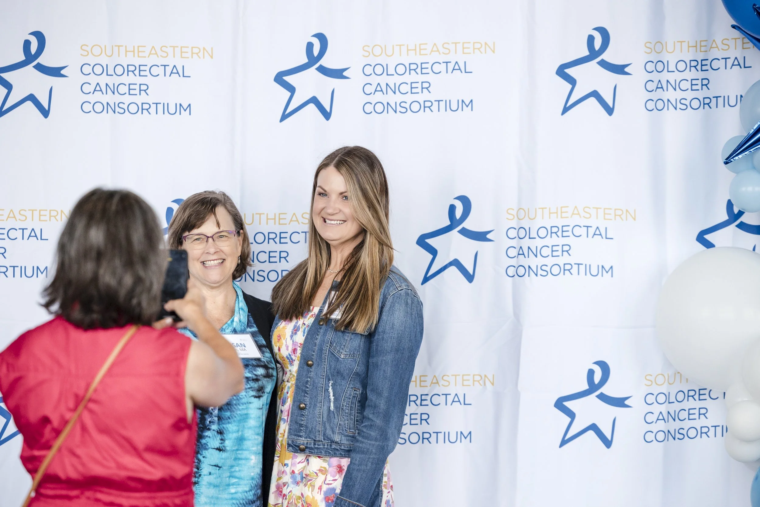 Two women posing for a photo at a Southeastern Colorectal Cancer Consortium event, with a volunteer taking their picture in front of a backdrop with the organization's logo and name, and balloons on the side.