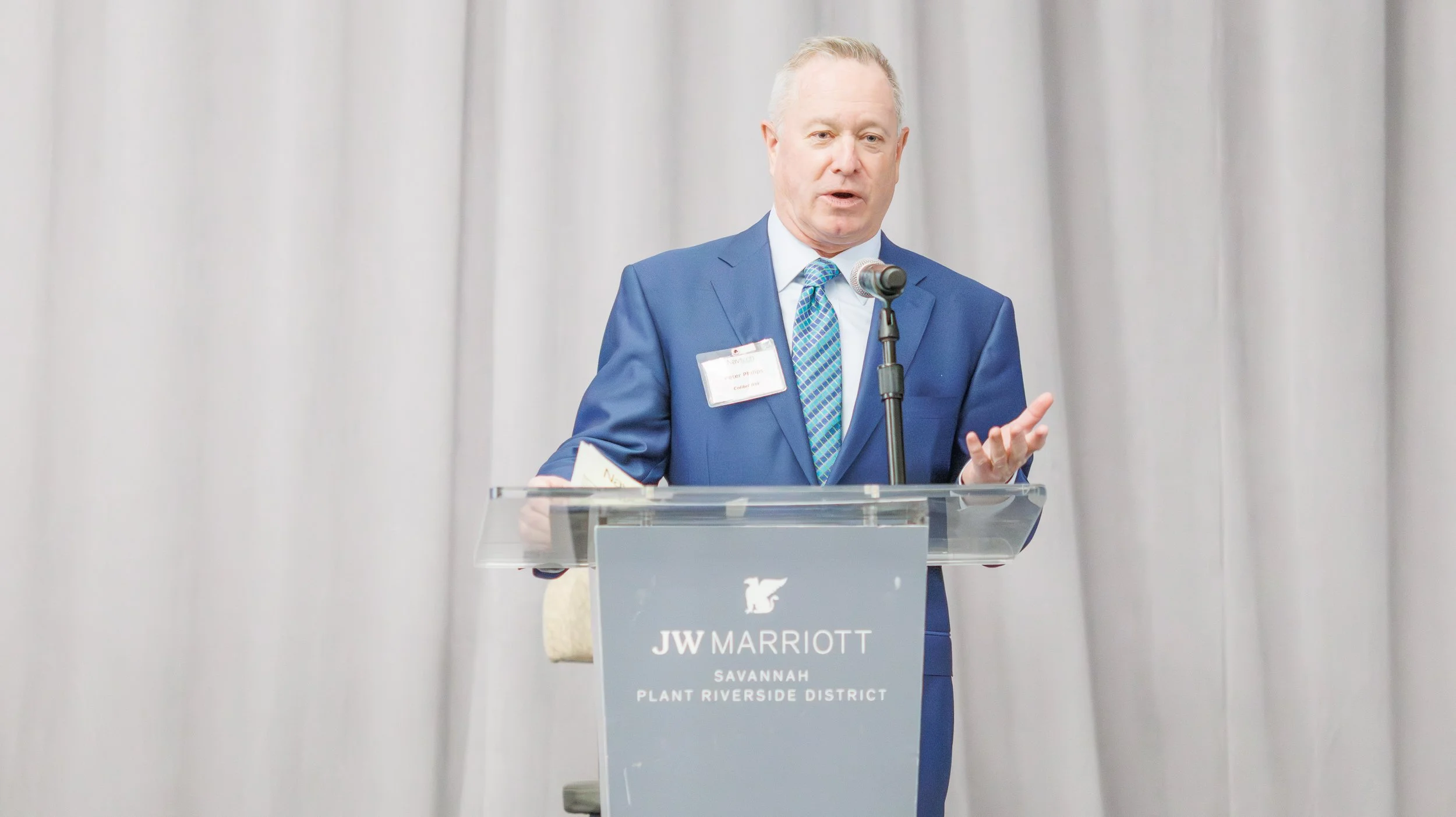 A man in a blue suit with a patterned tie is standing behind a clear lectern with a microphone, giving a speech or presentation. The background has plain light-colored curtains. The lectern has a sign that reads 'JW Marriott Savannah Plant Riverside 