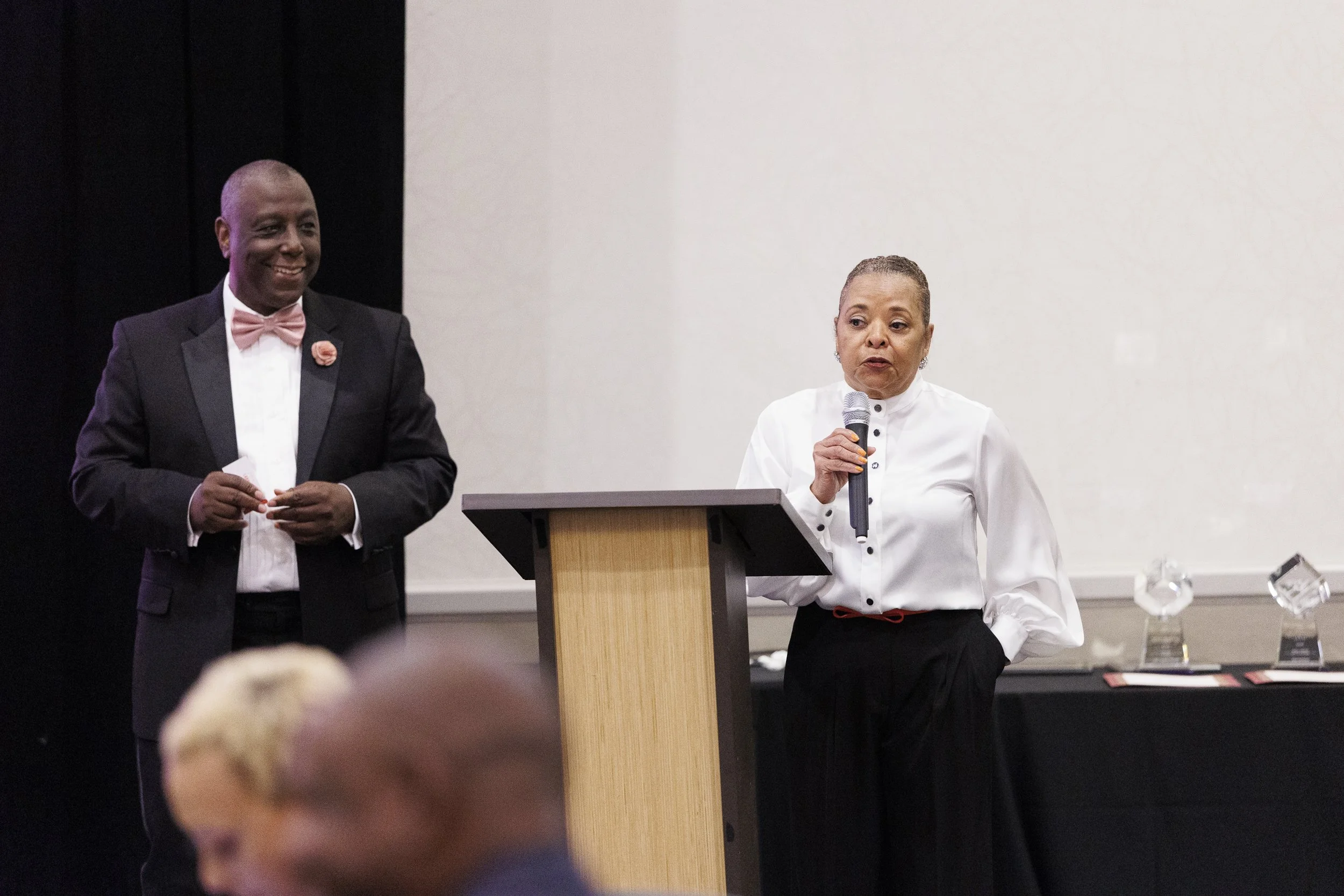 A woman in a white blouse speaking into a microphone at a podium, with a man in a black tuxedo and pink bow tie standing nearby and smiling.