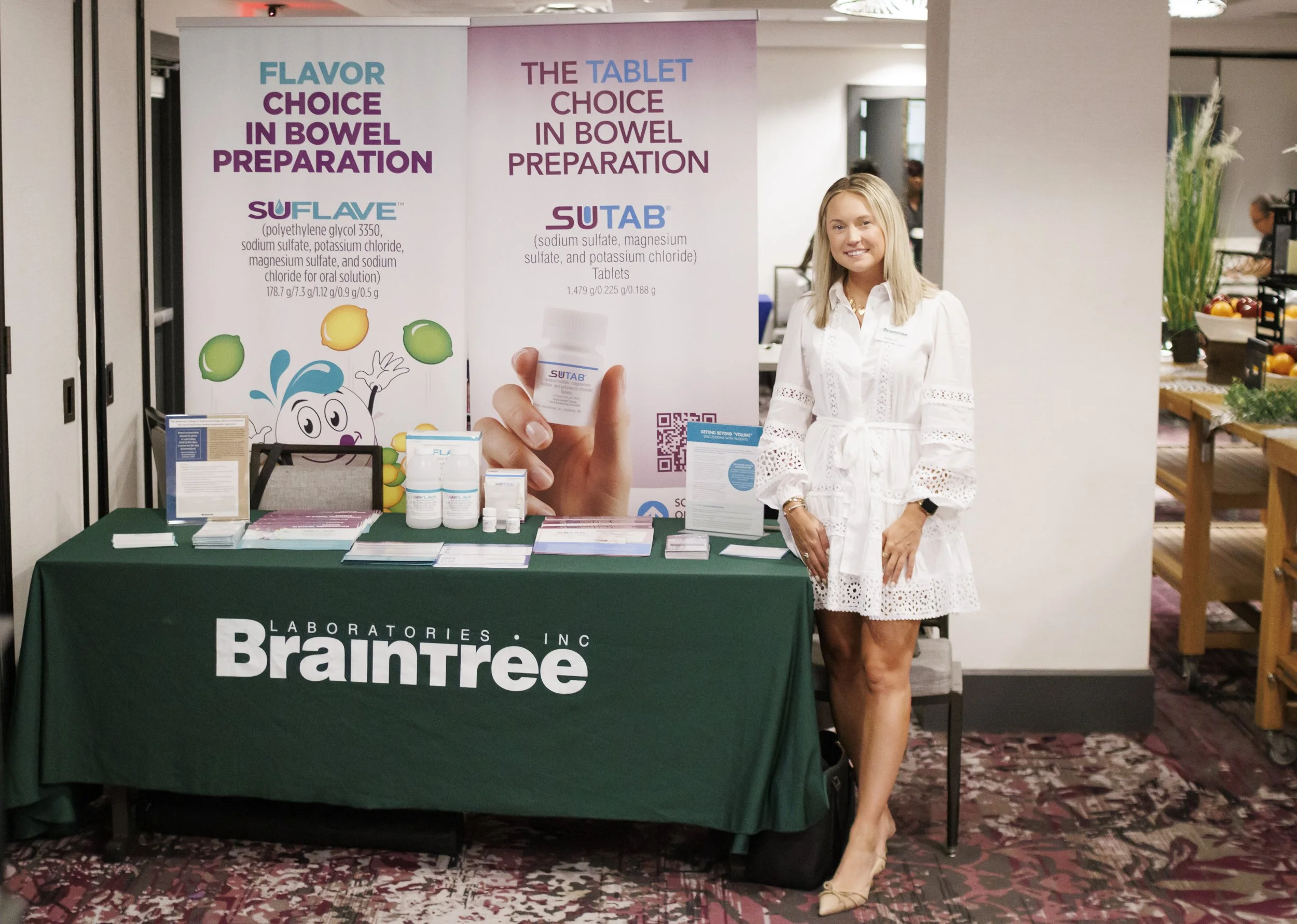 A woman in a white dress standing next to a table covered with a green tablecloth that has the Braintree Laboratories logo. The table displays informational brochures and medication bottles. Behind her are large banners promoting bowel preparation me