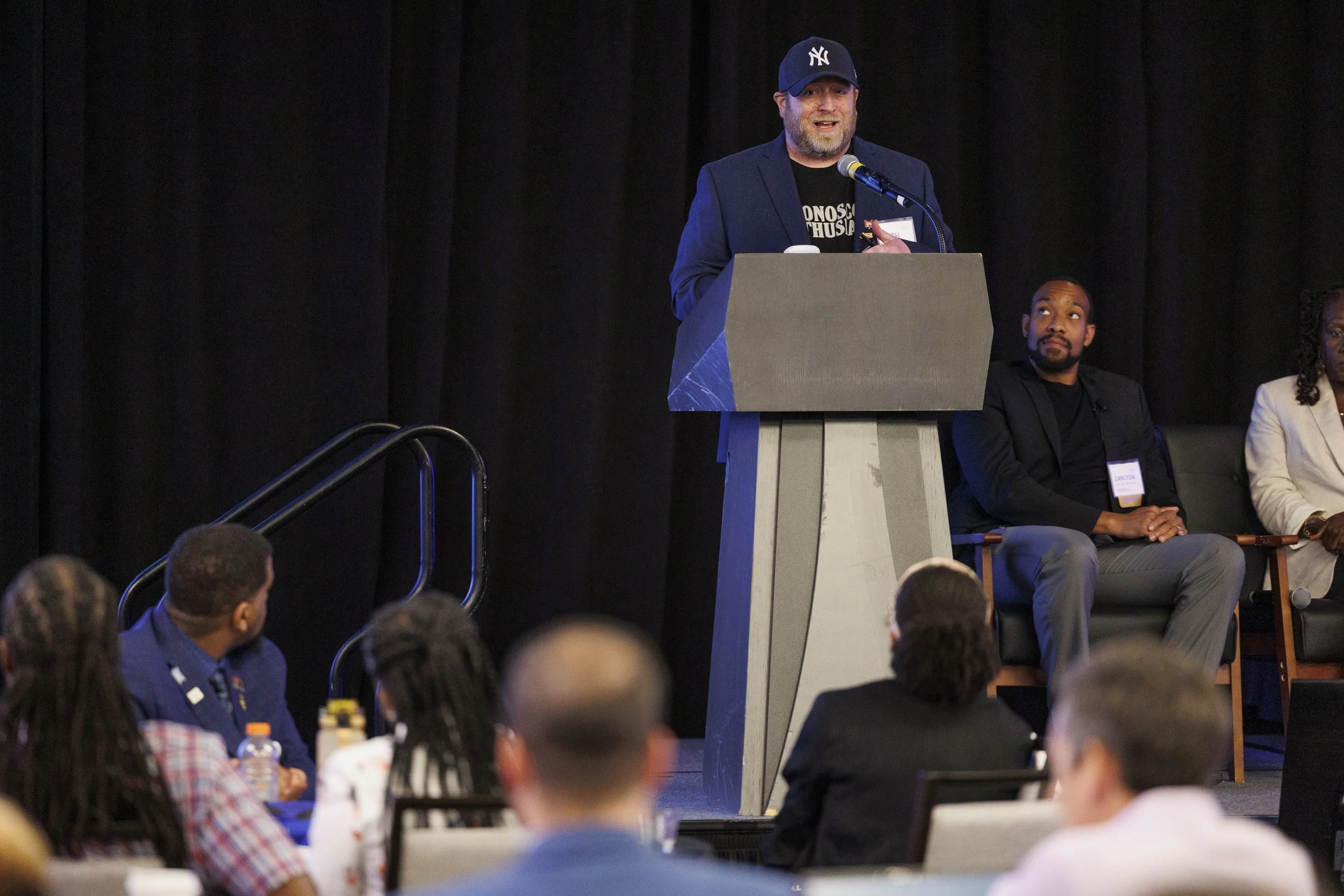 A man in a New York Yankees cap standing at a podium giving a speech at a conference, with three seated people behind him and an audience in front.
