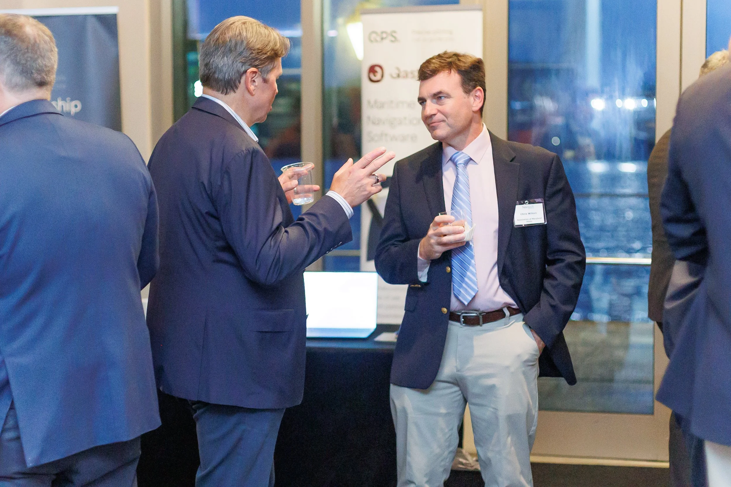 Group of men in business suits talking at a networking event, holding drinks, standing in front of a promotional display with a window behind them.