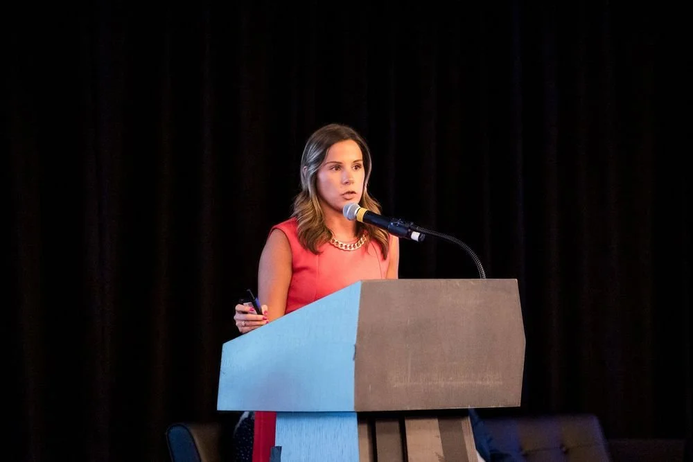 A woman in a red dress speaking at a podium with a microphone, on a stage with black curtains.