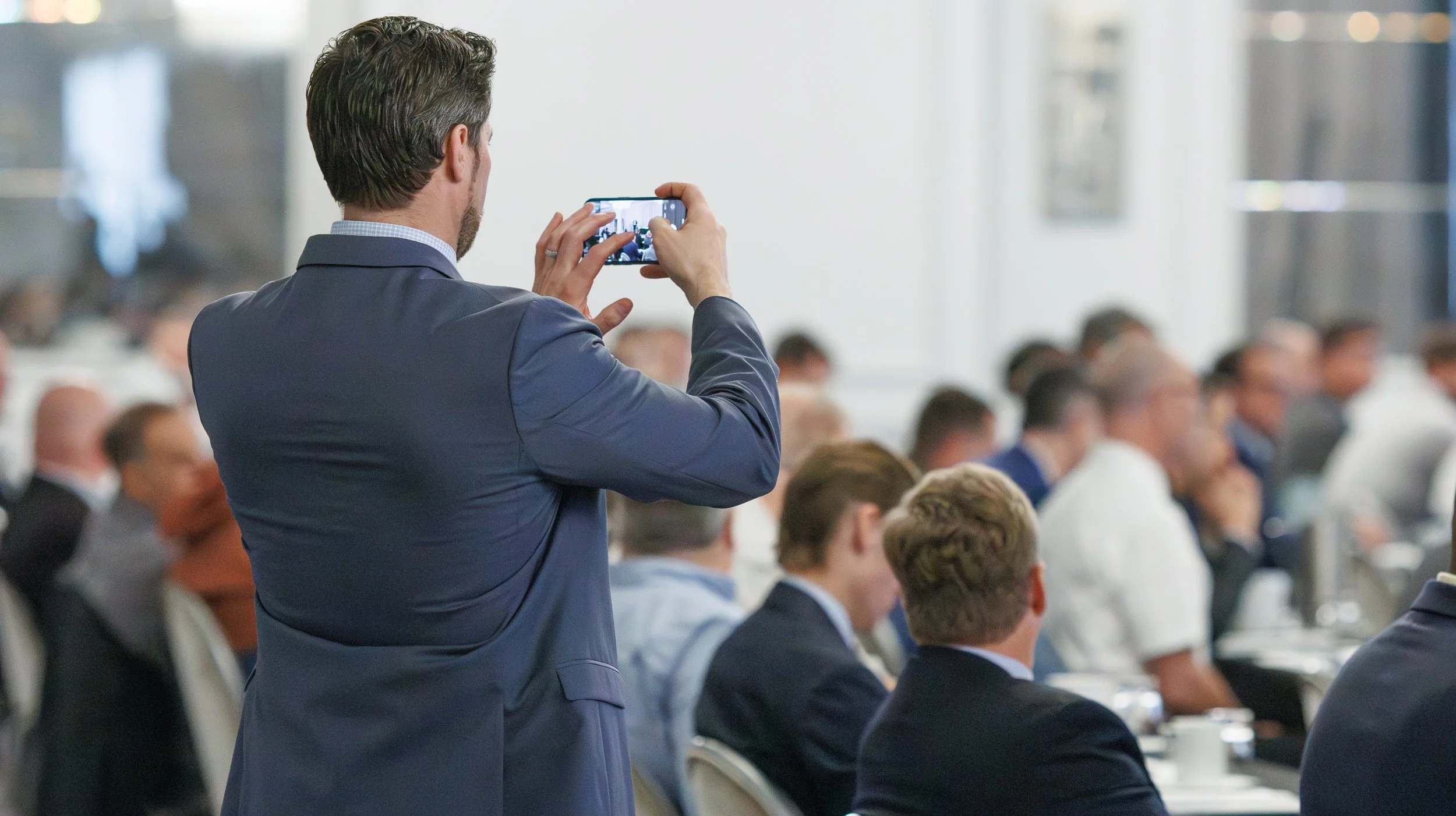 A man in a suit is taking a photo or video with his phone at a conference or seminar, with a group of people seated and listening.