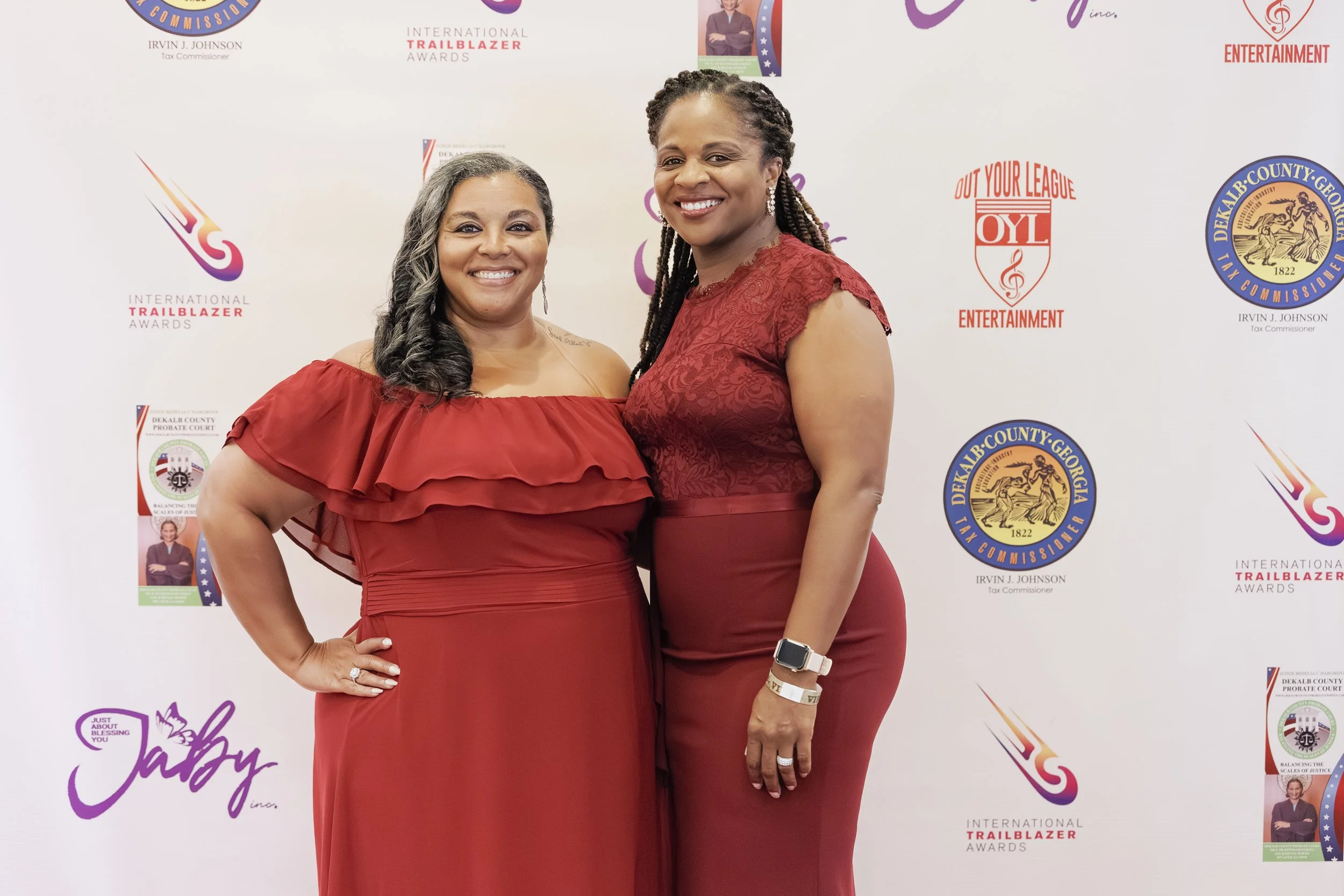 Two women in red dresses standing together, smiling at an event with a backdrop displaying various logos and text, including 'International Trailblazer Awards' and 'DeKalb County Georgia.'