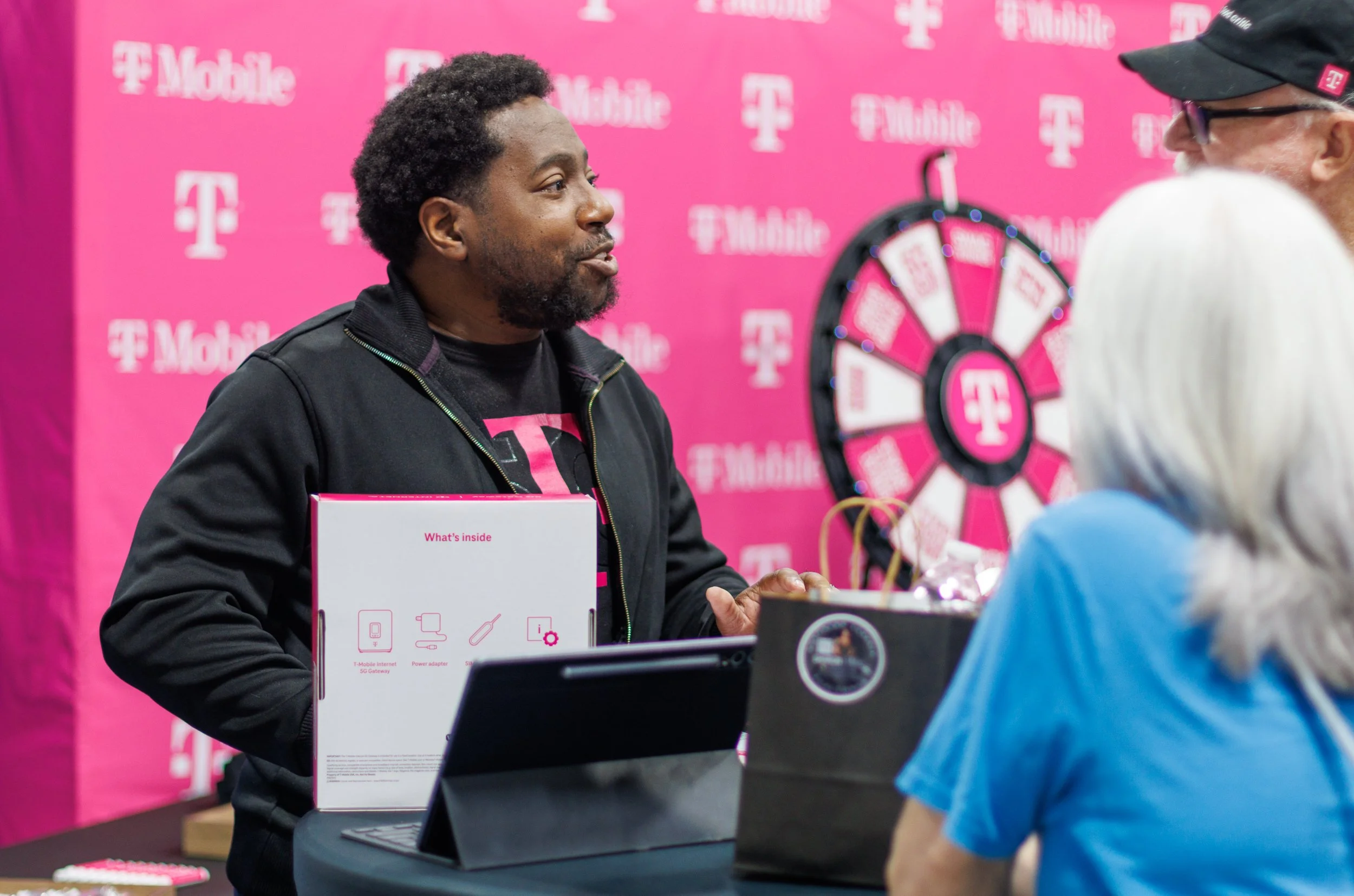 A man at a T-Mobile promotional booth is talking to two women and a man. The background is pink with T-Mobile logos, and there is a prize wheel. The man is wearing a black jacket and underneath a T-Mobile shirt. There are T-Mobile-branded items on th
