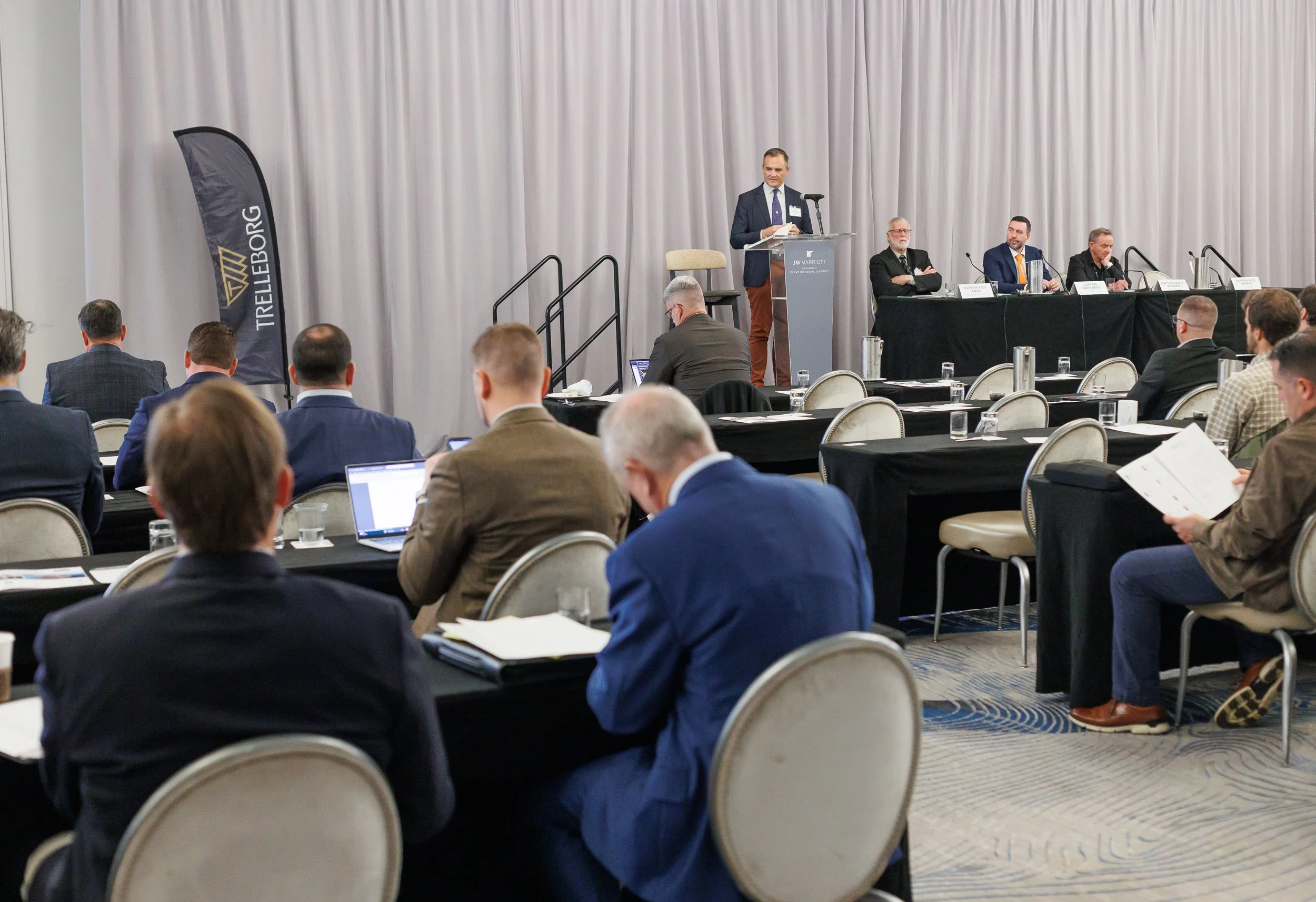 A professional conference with a speaker at a podium addressing an audience seated at tables, some using laptops.