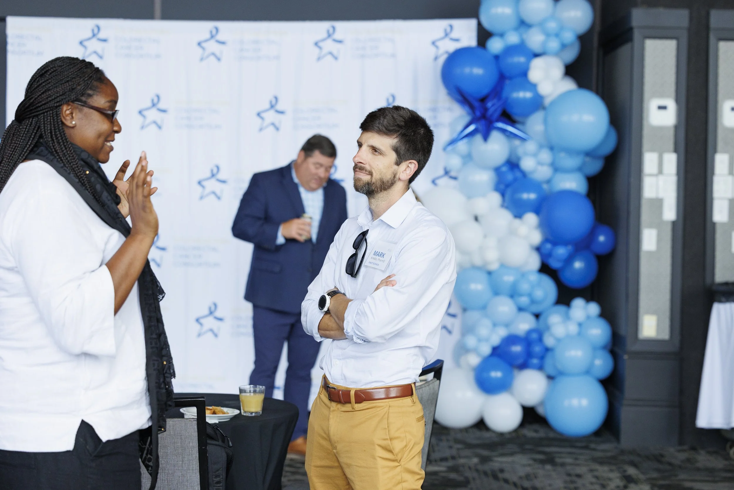 A woman with glasses and braids, wearing a white shirt, is speaking to a man with a beard, sunglasses hanging from his shirt, dressed in a white shirt and khaki pants, has his arms cross and is listening attentively at a corporate event with blue and