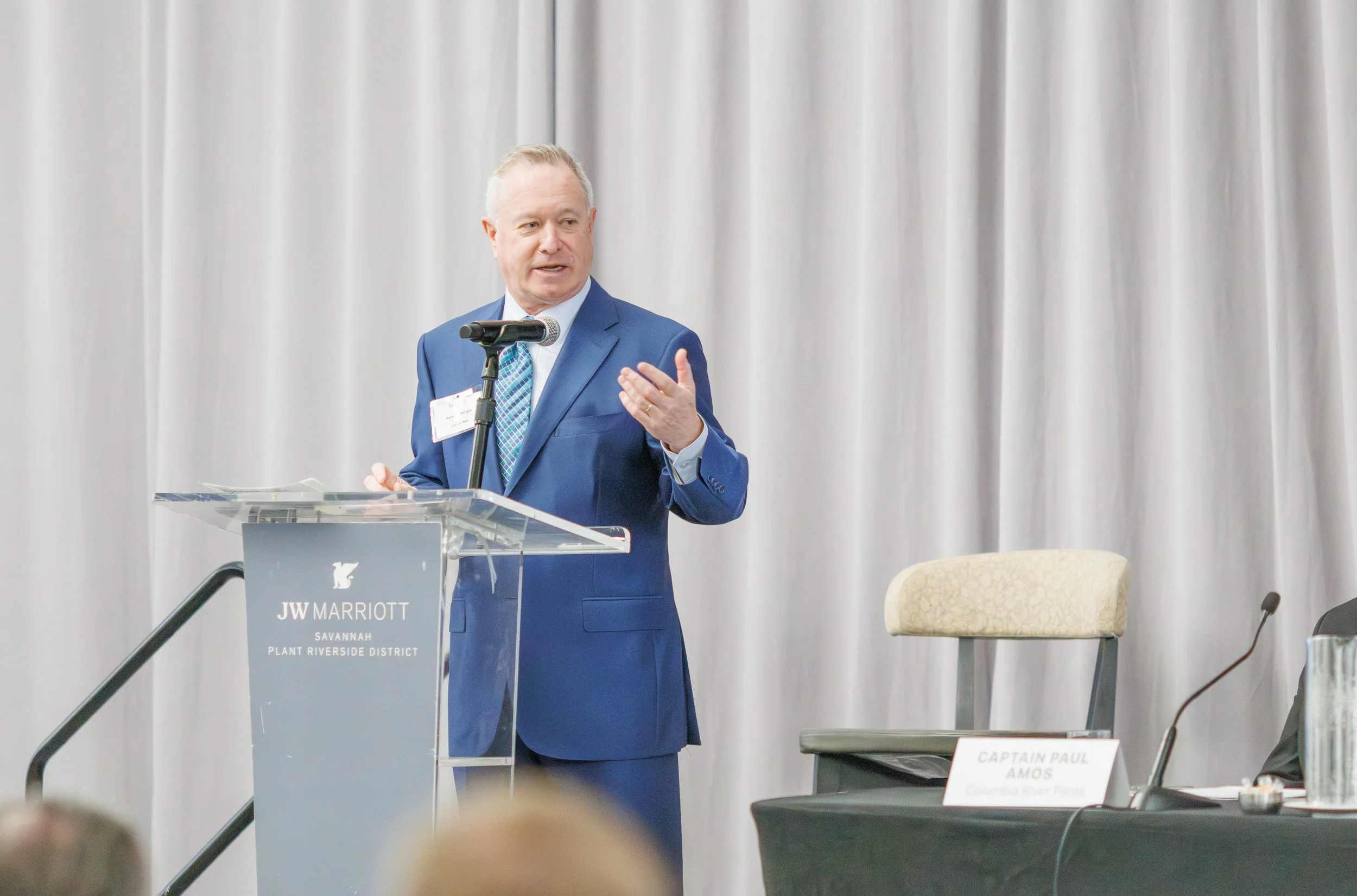 A man in a blue suit speaking at a podium during a conference at JW Marriott Savannah Plant Riverside District. A sign on the podium reads 'JW Marriott Savannah Plant Riverside District'. There are curtains in the background and a nameplate for Capta