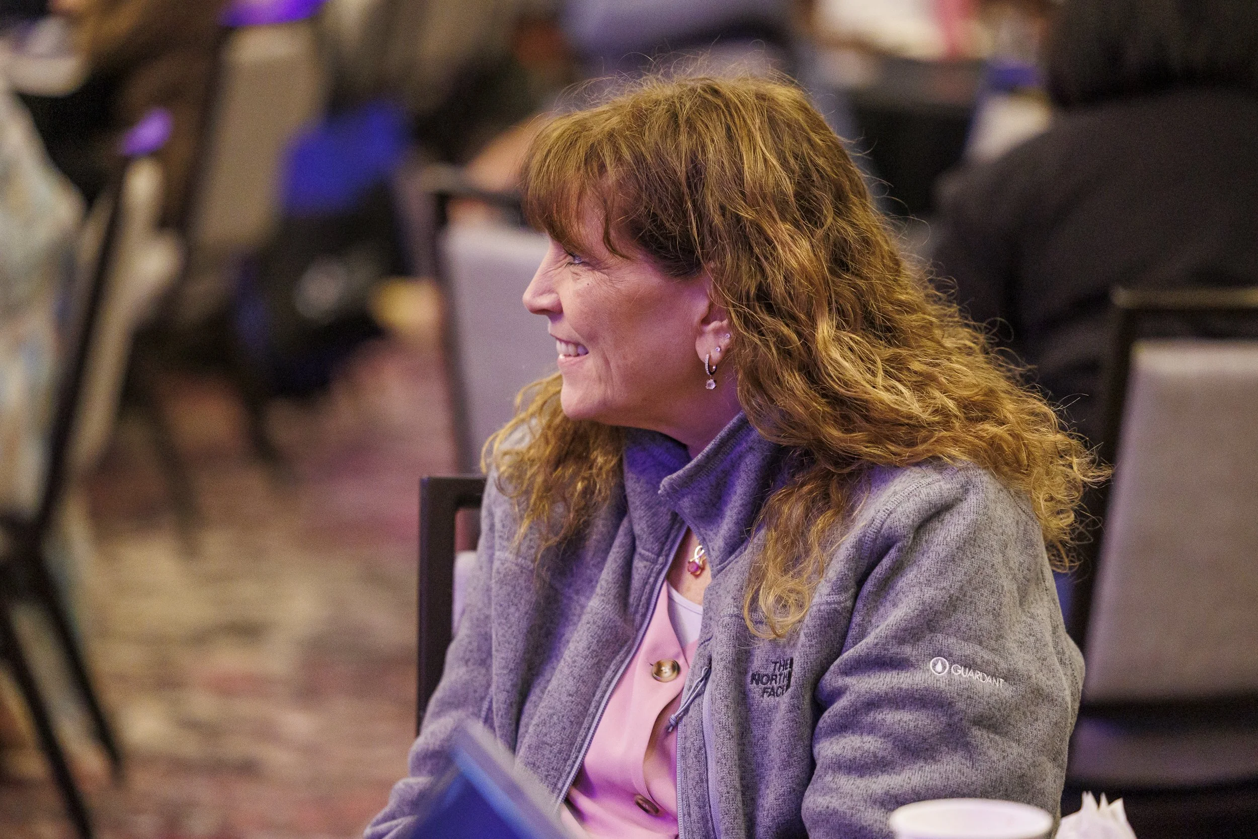 Smiling woman with curly red hair wearing earrings and a gray jacket, sitting at an event or conference.