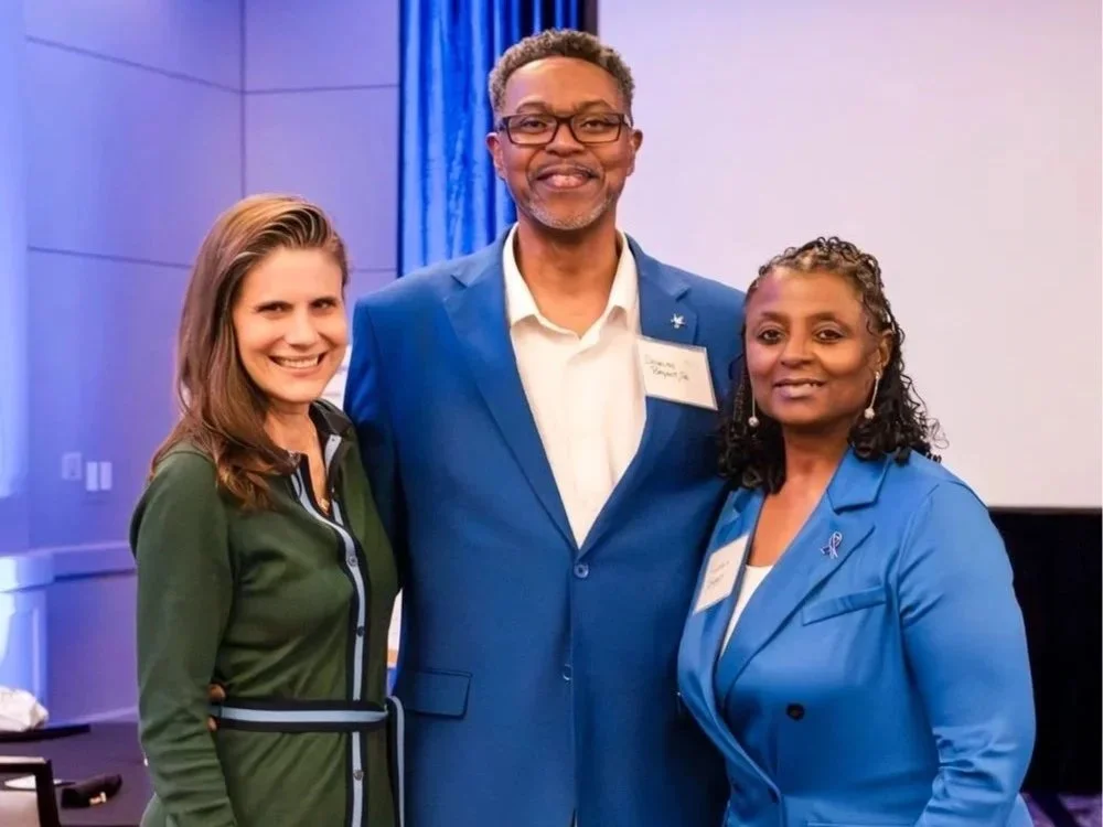 Three people standing together at an indoor event, smiling for the camera. The man in the middle is wearing glasses and a blue suit, with a name tag. The woman on the left has long brown hair and is wearing a dark green jacket with black and white de