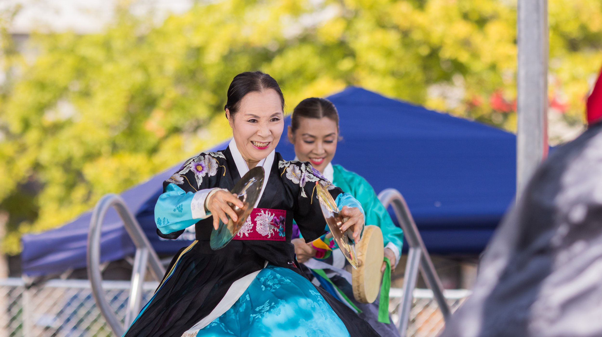 Women in traditional Korean hanbok costumes smiling and holding shoes outdoors with yellow and green trees in the background.