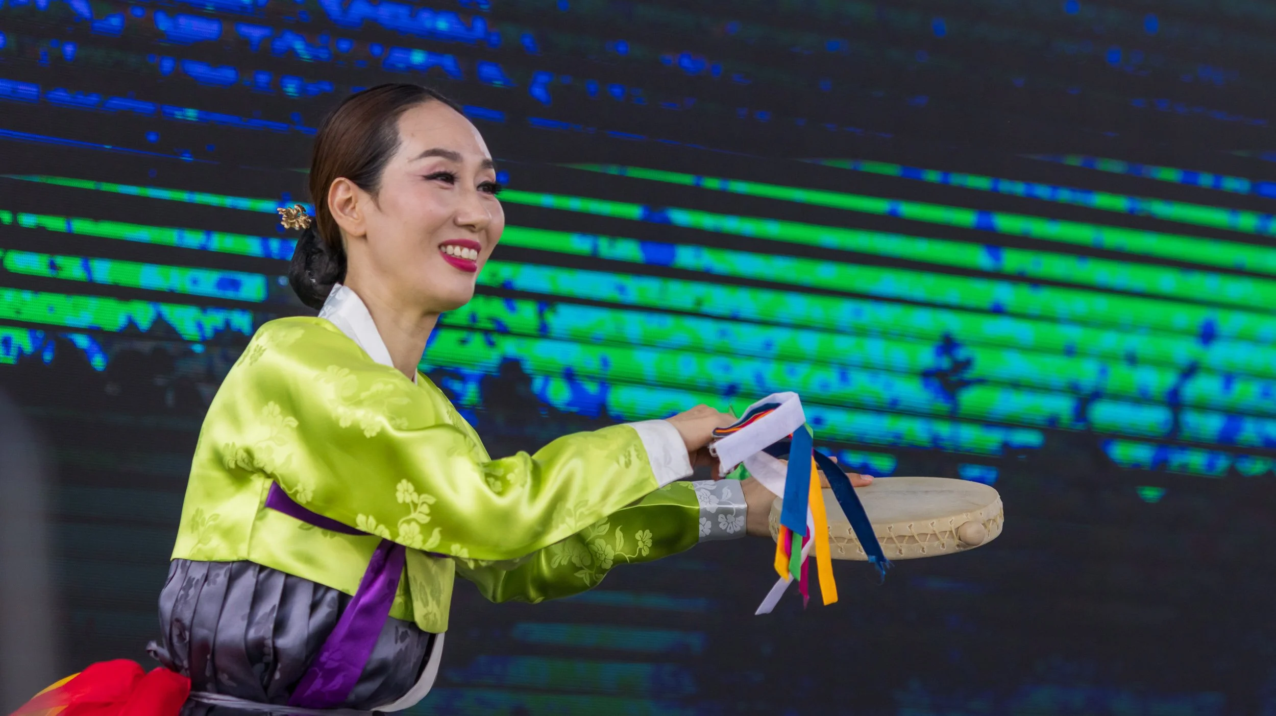 A woman in a traditional Korean dress (hanbok) smiling and participating in a cultural event or celebration, with a background of colorful digital or illuminated display.