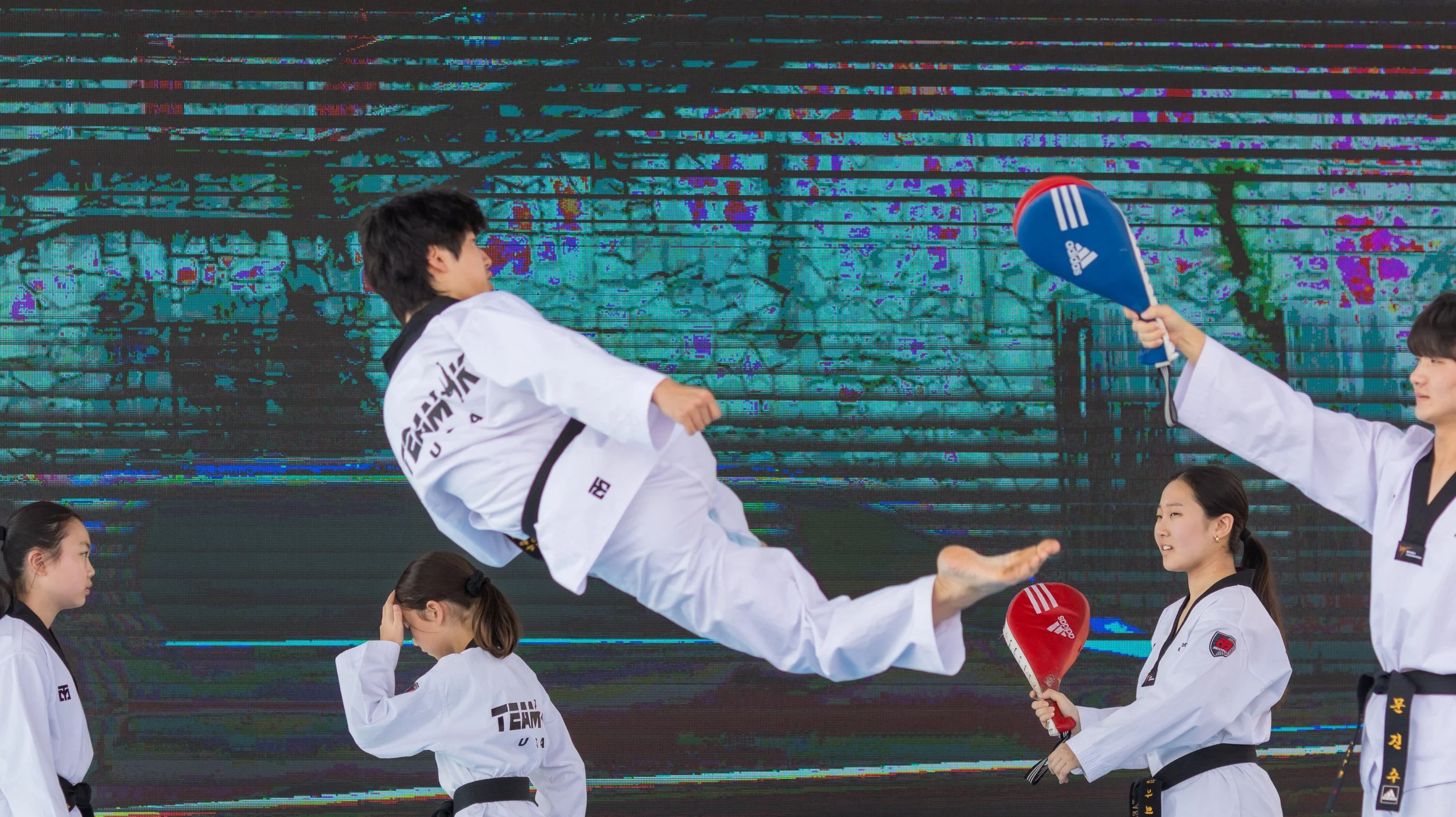 Group of martial artists practicing taekwondo in uniform, with one person kicking and another holding a paddle target, in front of an LED background.
