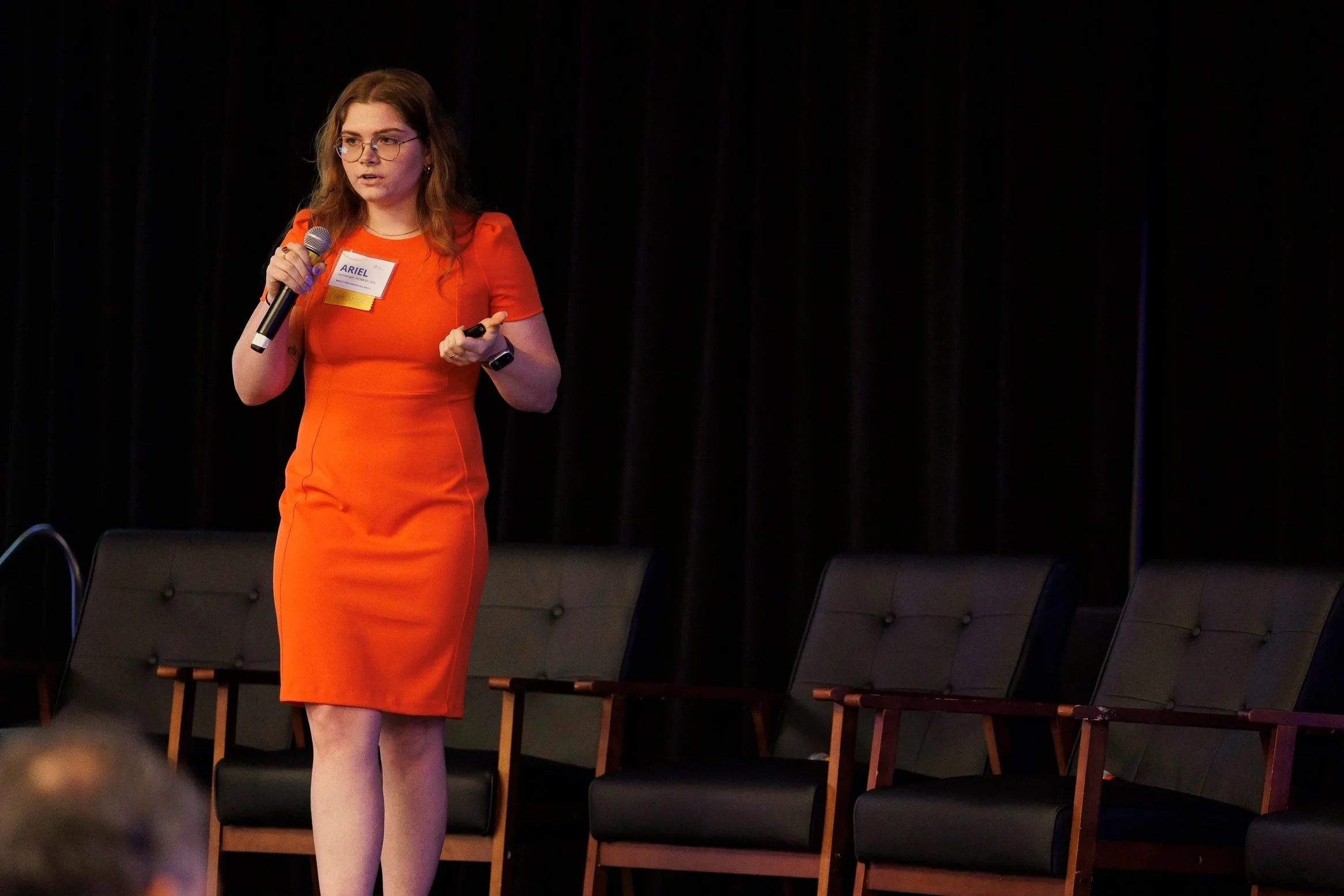 A woman in a red dress holding a microphone and a presentation remote on stage, with a black curtain background and empty chairs behind her.
