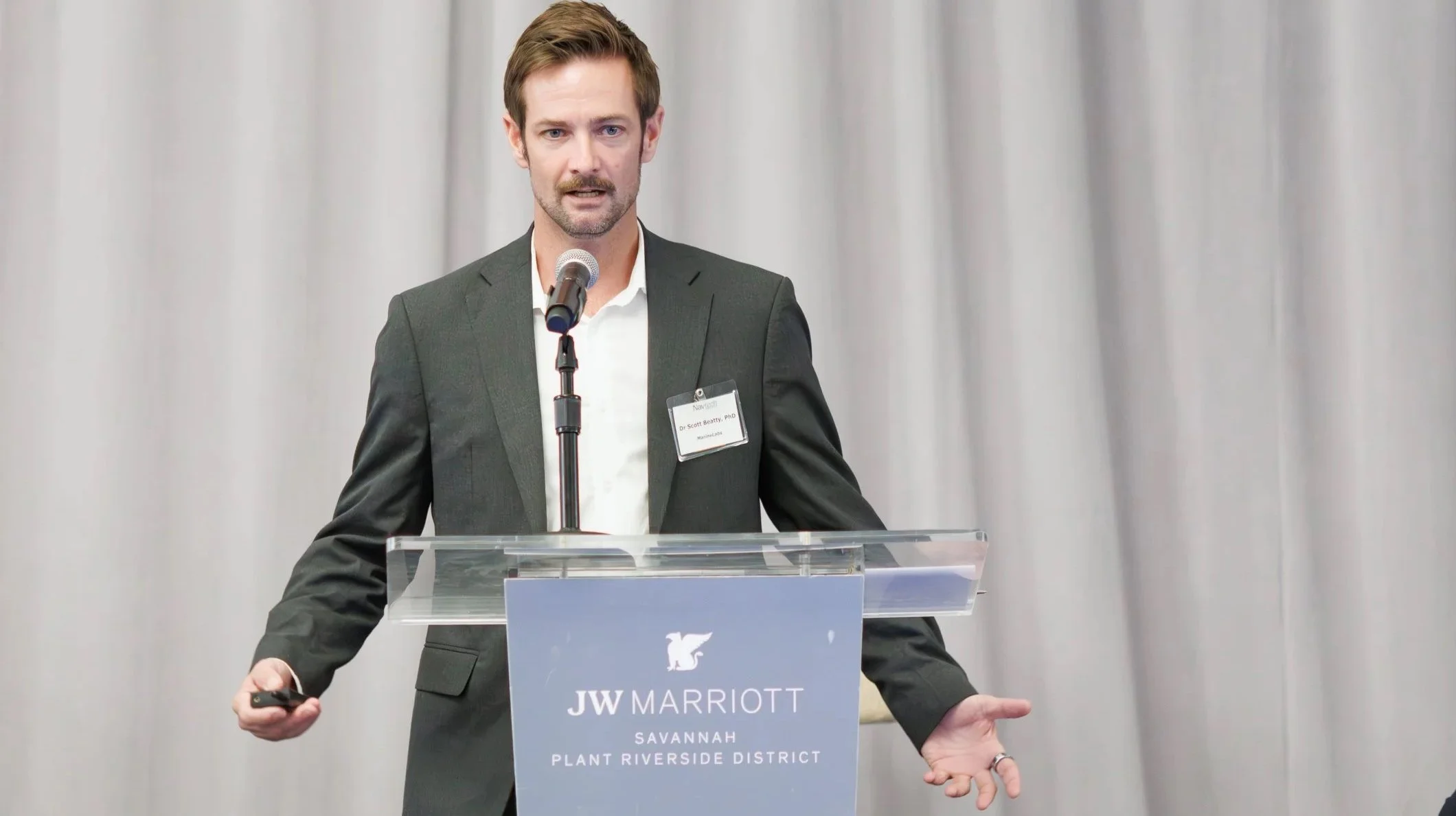 A man in a dark gray suit is standing at a podium with a microphone, giving a speech at a conference. The podium has a sign that reads 'JW Marriott Savannah Plant Riverside District.' The man has short brown hair and a beard, and is wearing a name ba