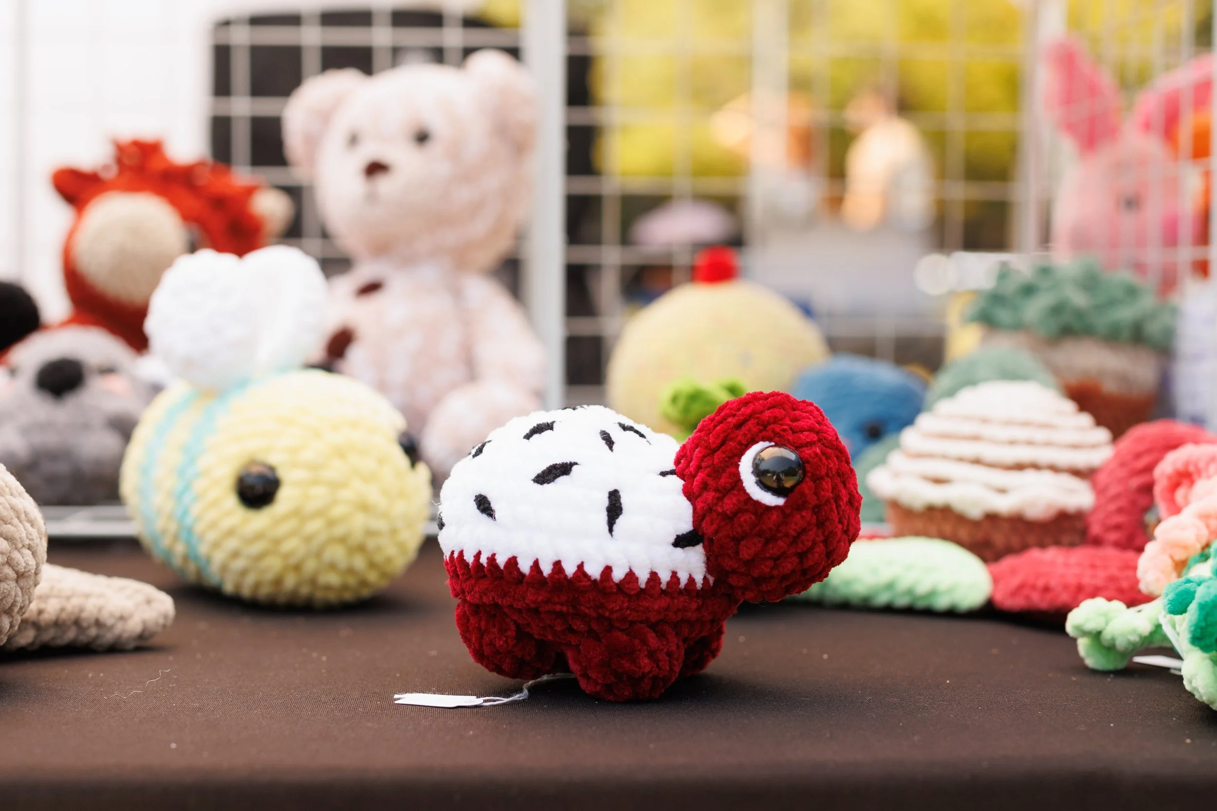 Colorful crochet or knitted animal plush toys, including a red and white sheep, displayed on a table at an outdoor market or craft fair.