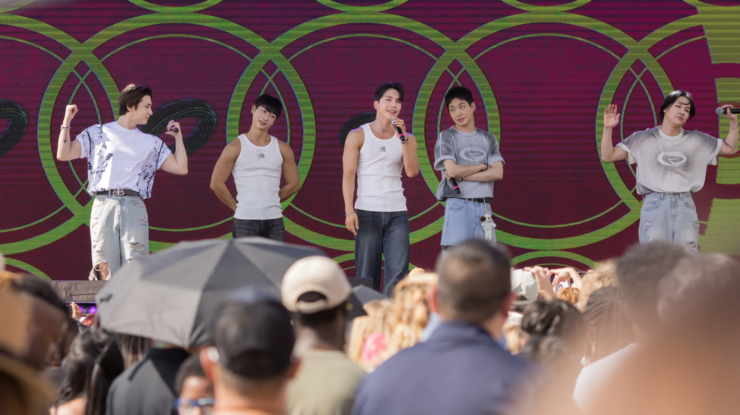 Six young men performing on an outdoor stage with a colorful, geometric background. They are dressed casually in sleeveless shirts and jeans, facing the audience with some holding microphones.