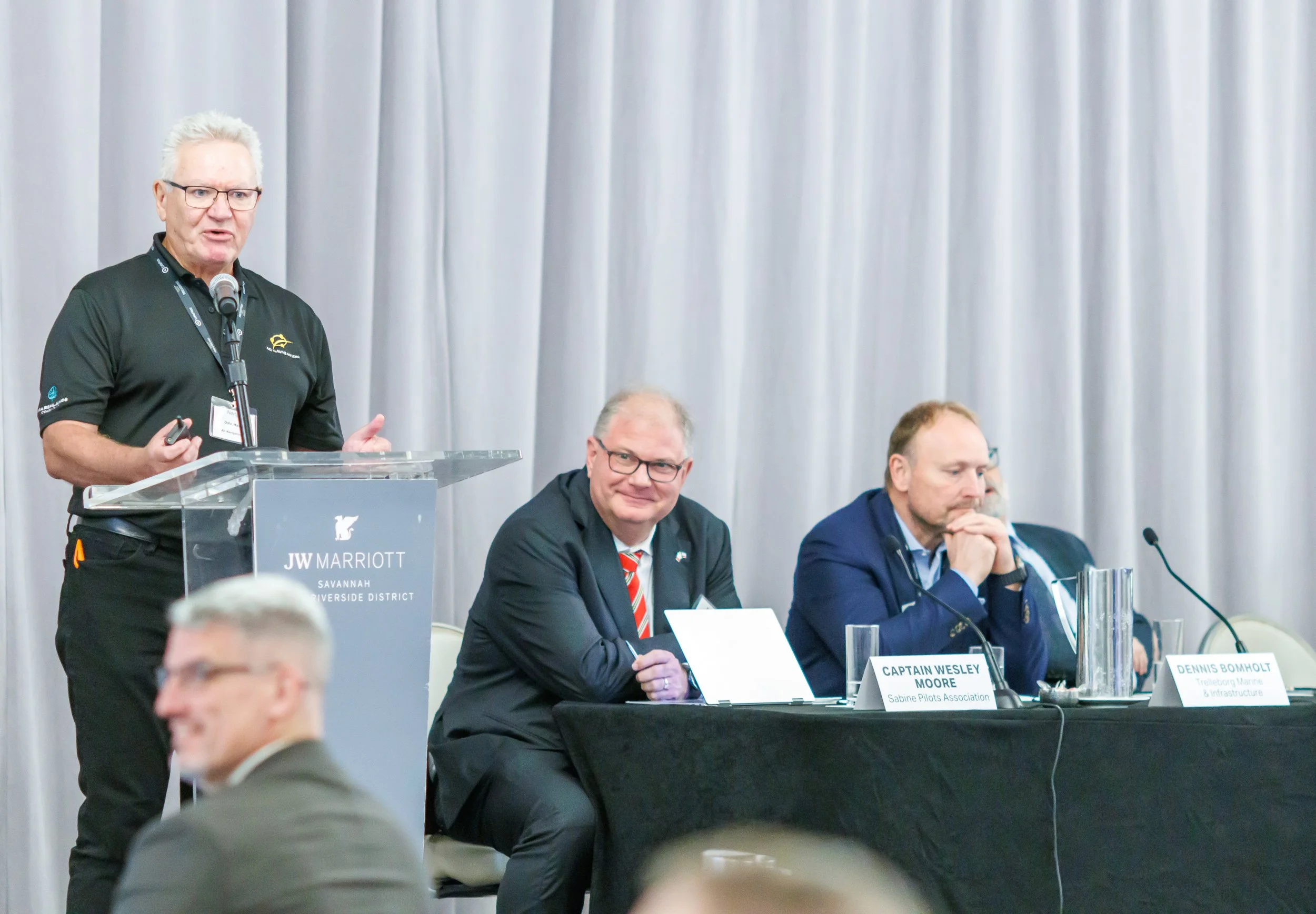 A man with gray hair and glasses is speaking at a podium with a microphone, wearing a black polo shirt with a logo, at a conference at JW Marriott in Savannah. Three other men are seated at a table beside him, with nameplates in front of them. The ma