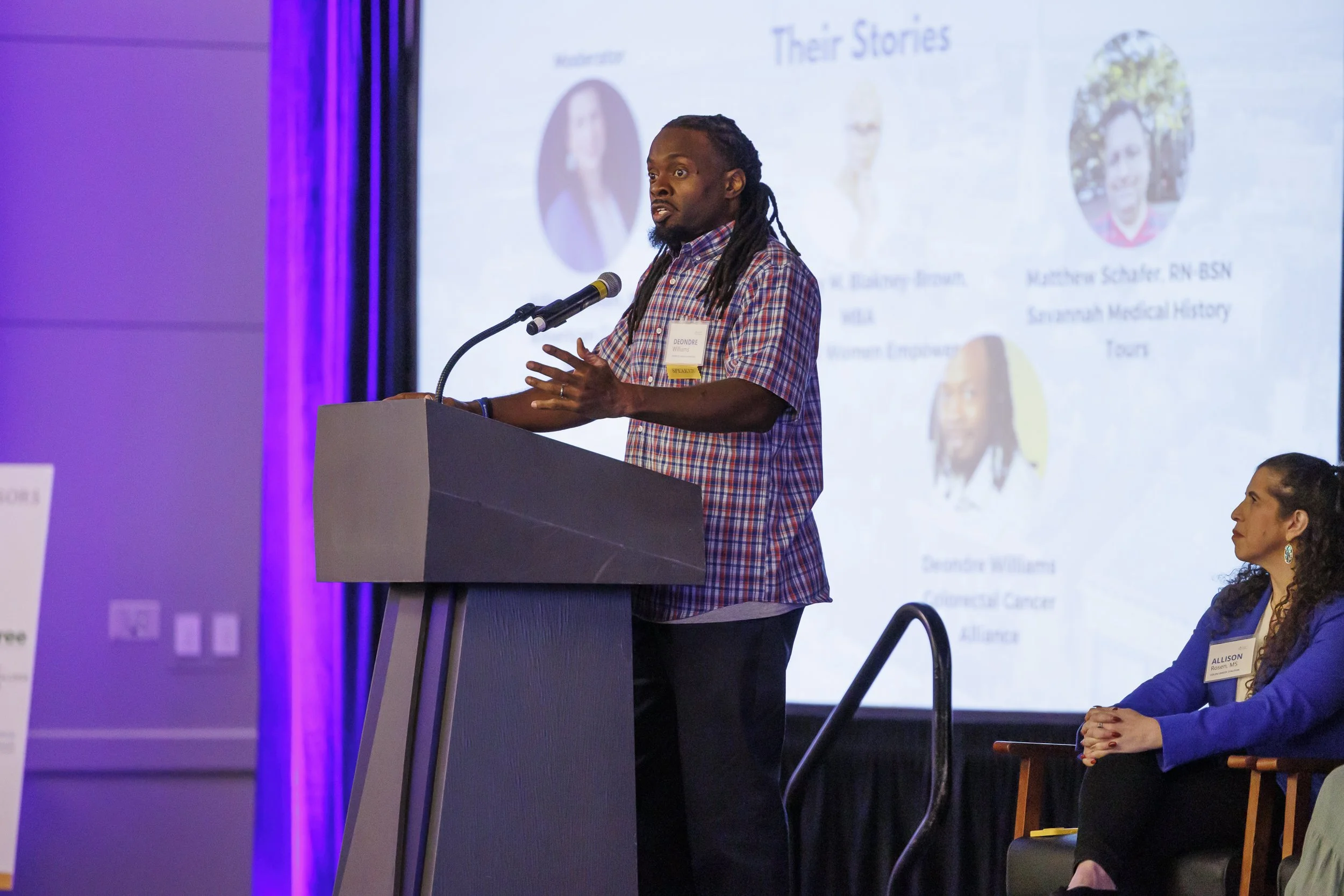 A man with long dreadlocks speaking at a podium during a conference, with a woman seated nearby. Behind them is a large screen displaying slides with photos and text titled 'Their Stories'.