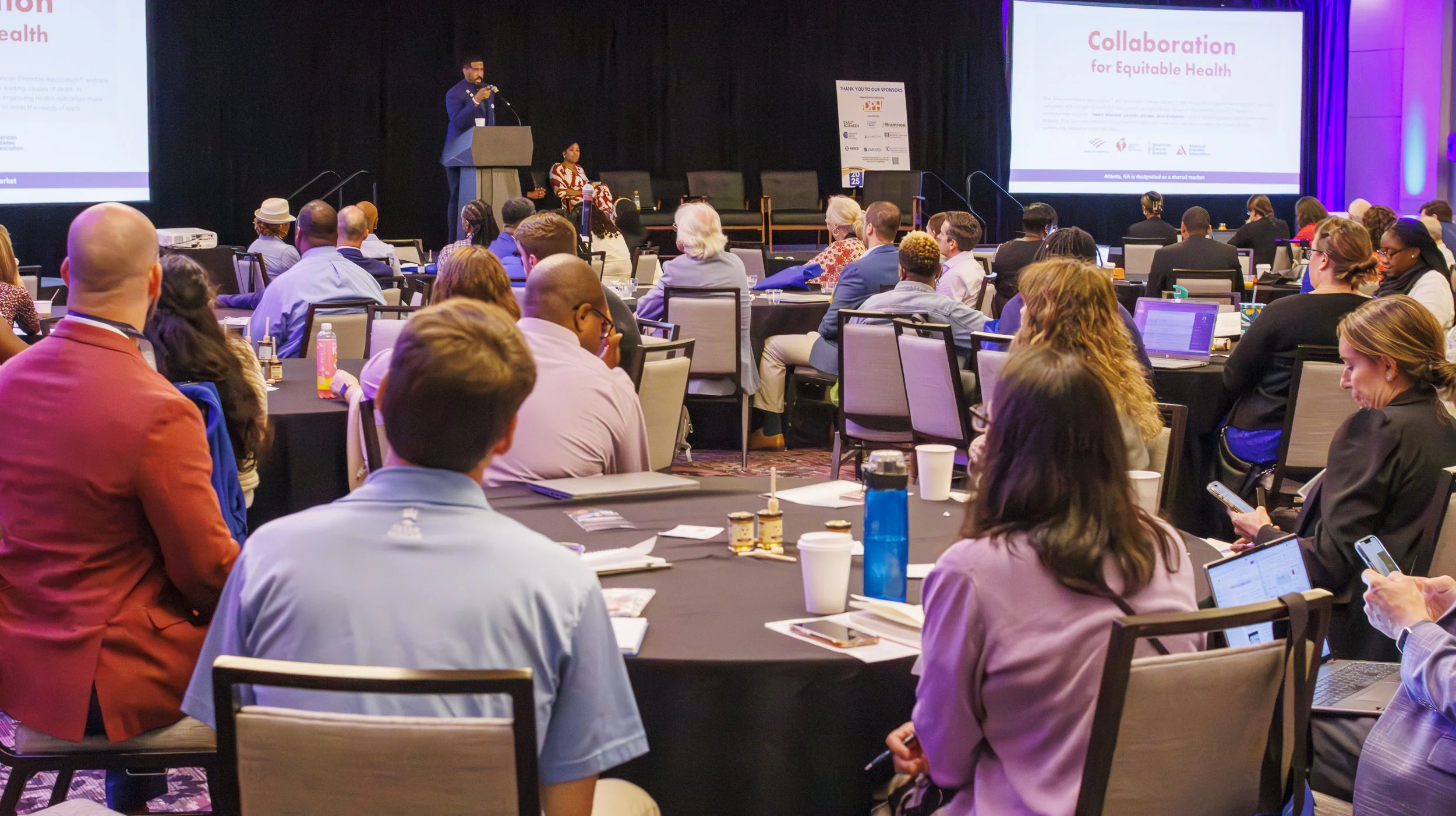 Conference room with attendees seated at round tables, listening to a speaker on stage and a presentation slide that reads 'Collaboration for Equitable Health'.