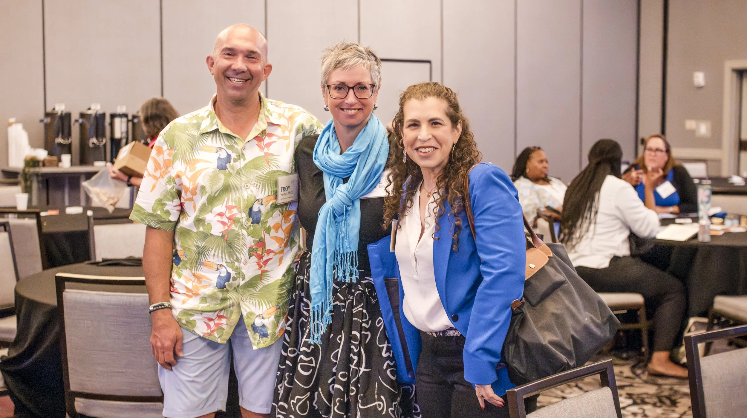 Three people standing together smiling at a conference, with women sitting at tables in the background.