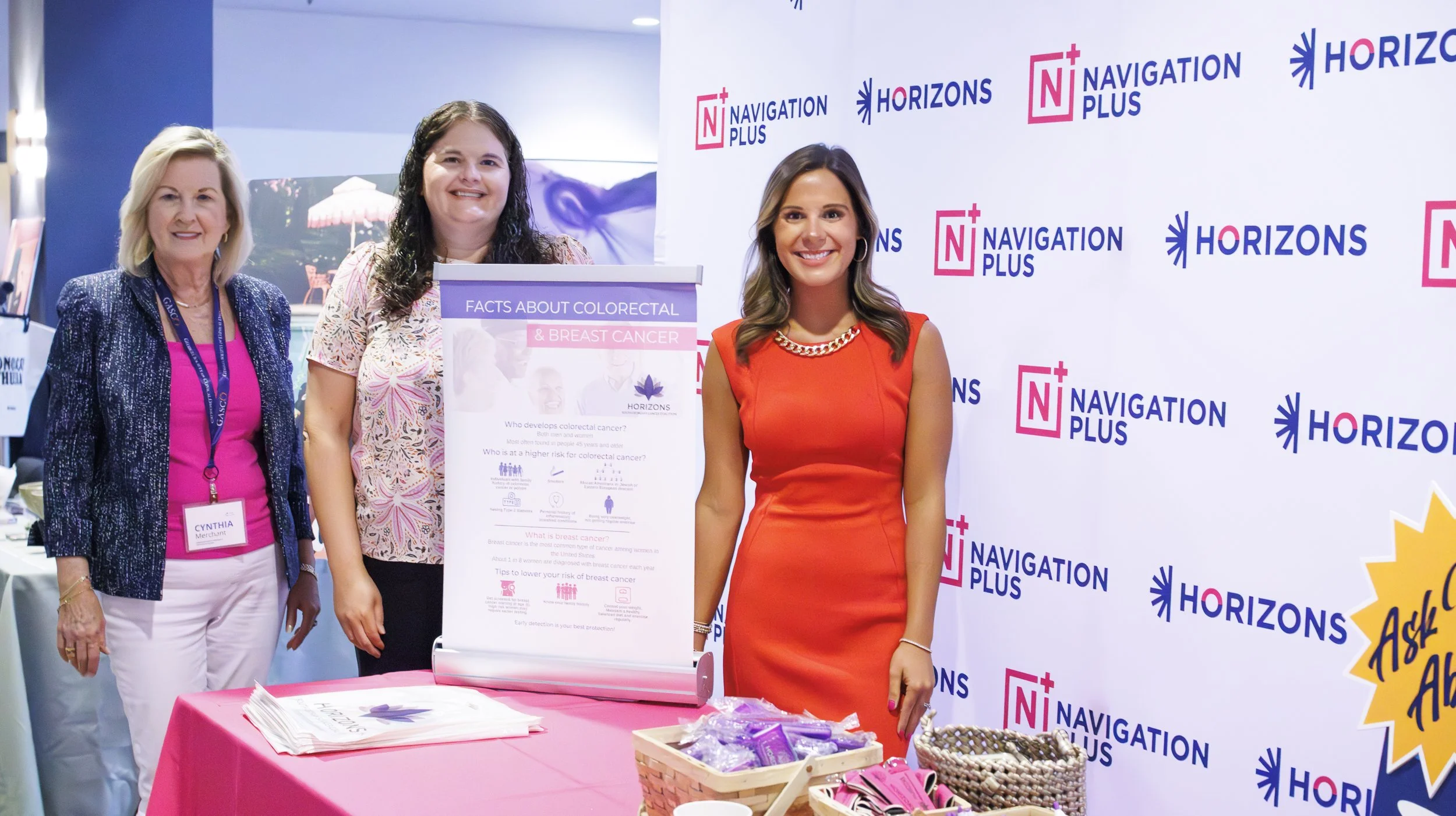 Three women standing in front of a promotional backdrop at a health awareness event. One woman is wearing a pink top and white pants with a name badge, the second woman is holding a poster about colorectal and breast cancer, and the third woman is in