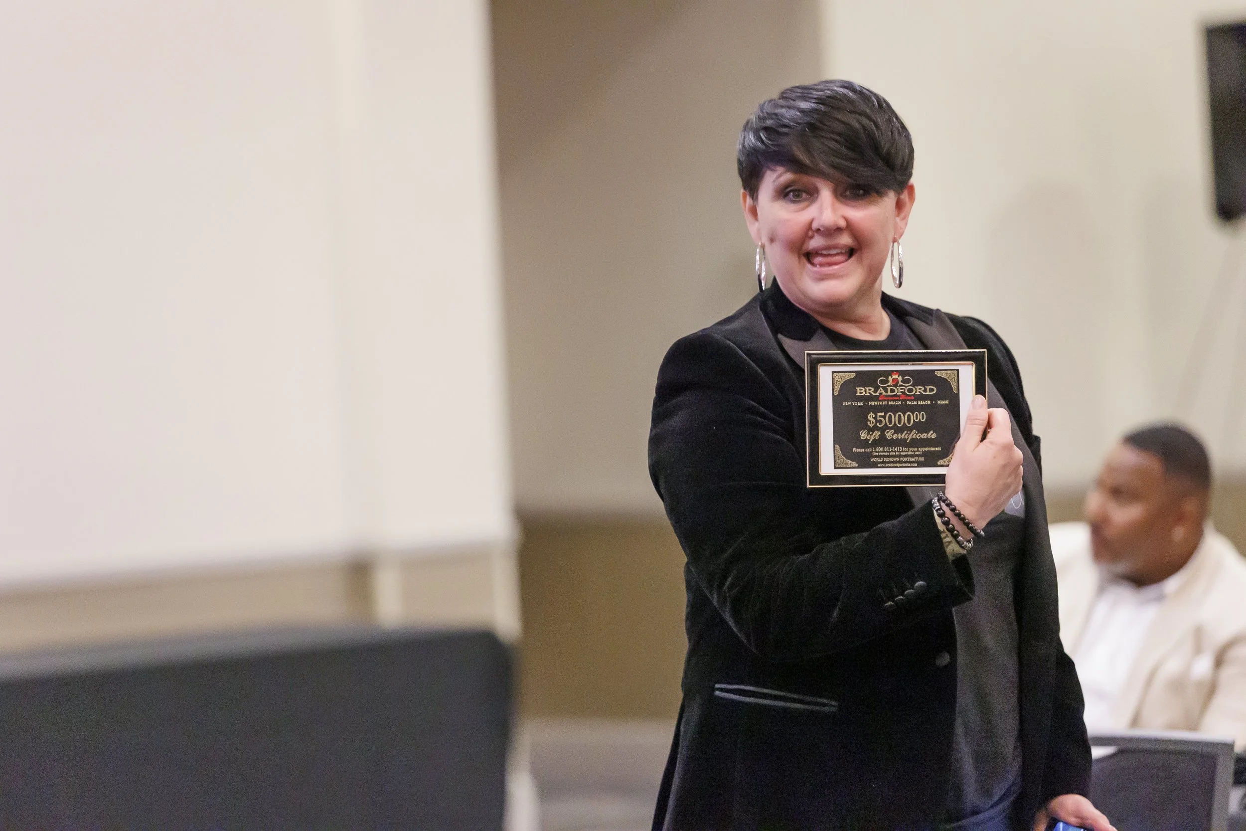 A woman with short dark hair and hoop earrings is holding a large gift certificate and speaking at an indoor event. The certificate is for 5 million dollars from Bradford.