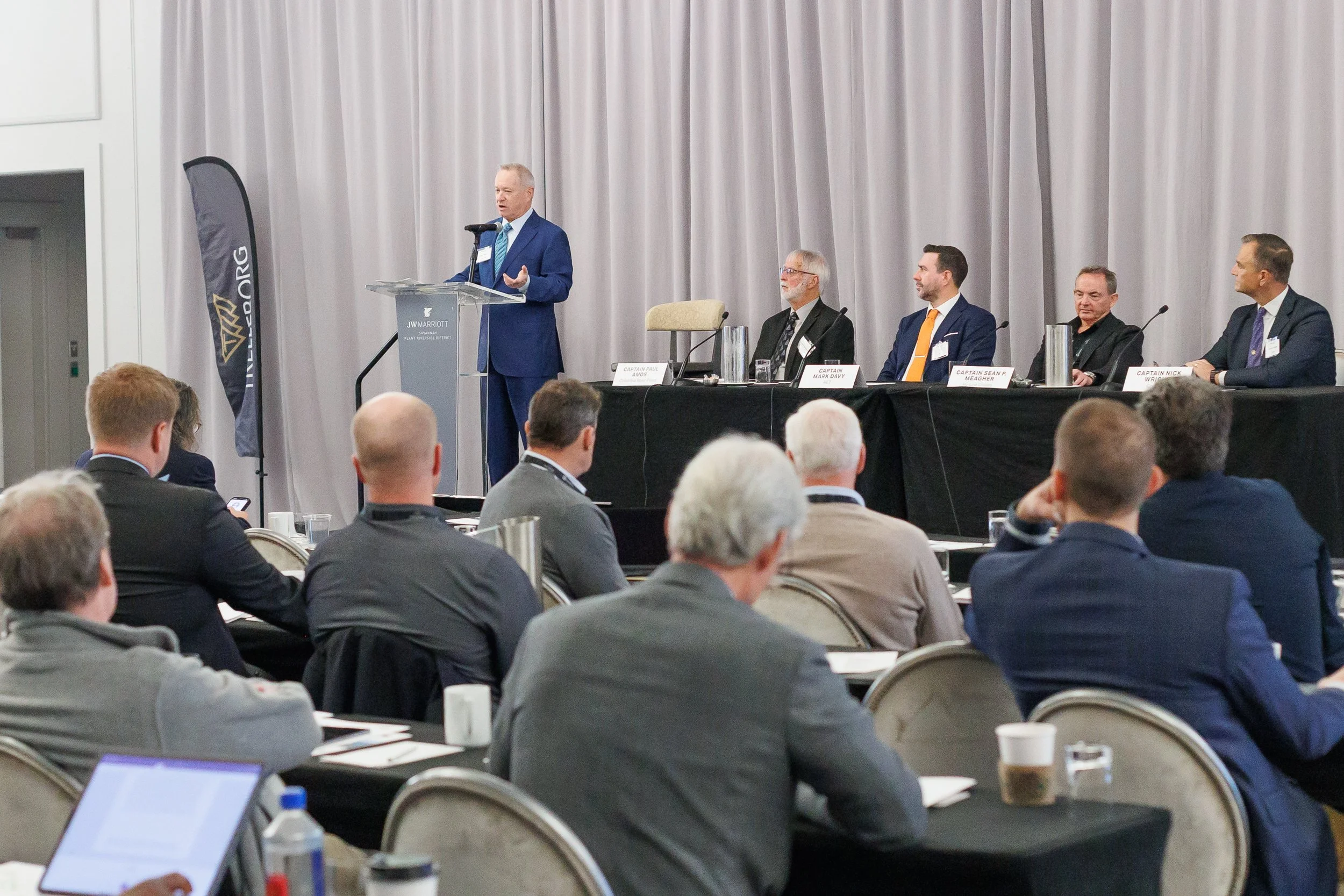 A man in a blue suit giving a speech at a conference, with five panelists seated behind him at a long table, in front of an audience in a large conference room.