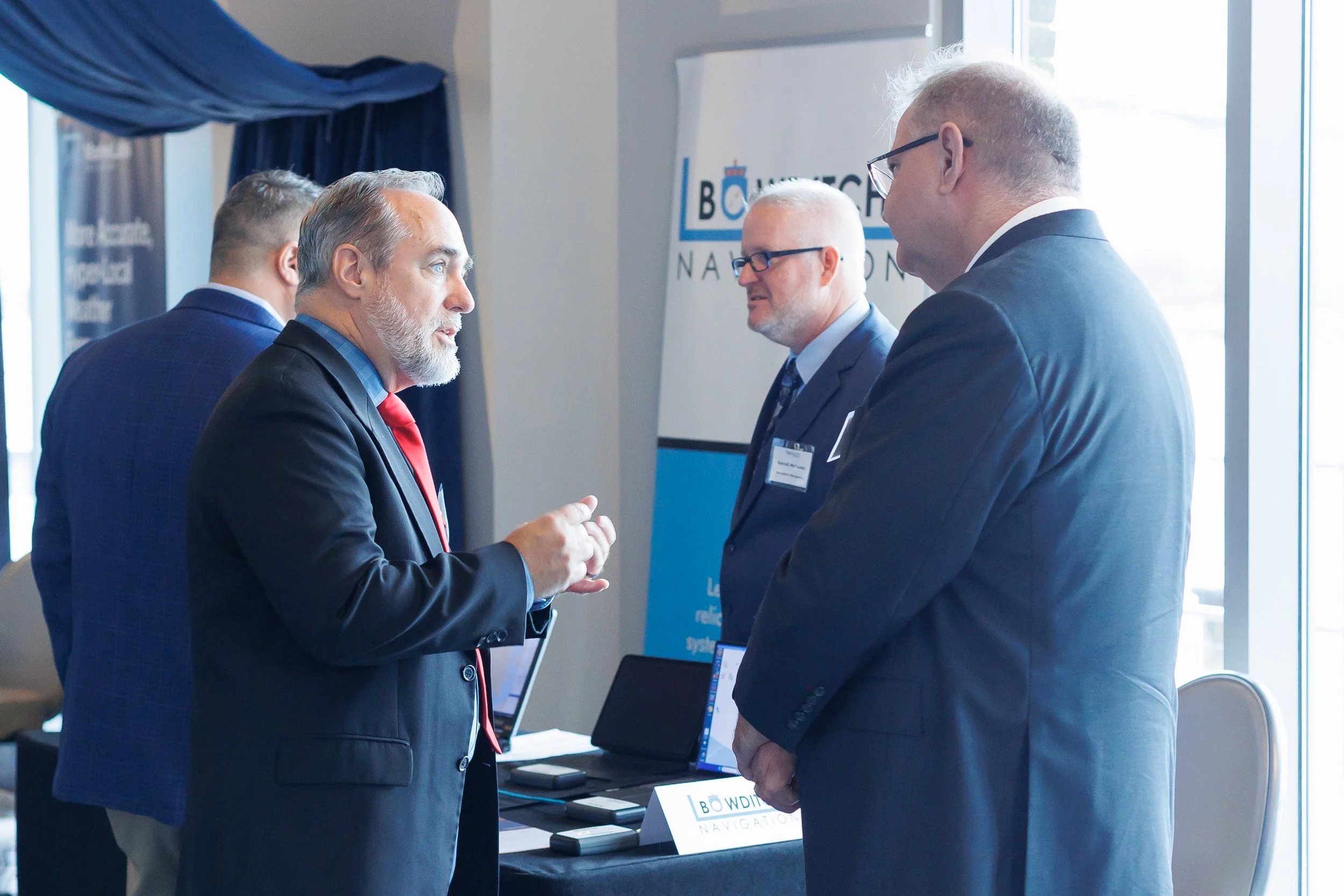 Four men in business suits engaged in conversation at a professional conference, with a booth setup behind them and a large window nearby.