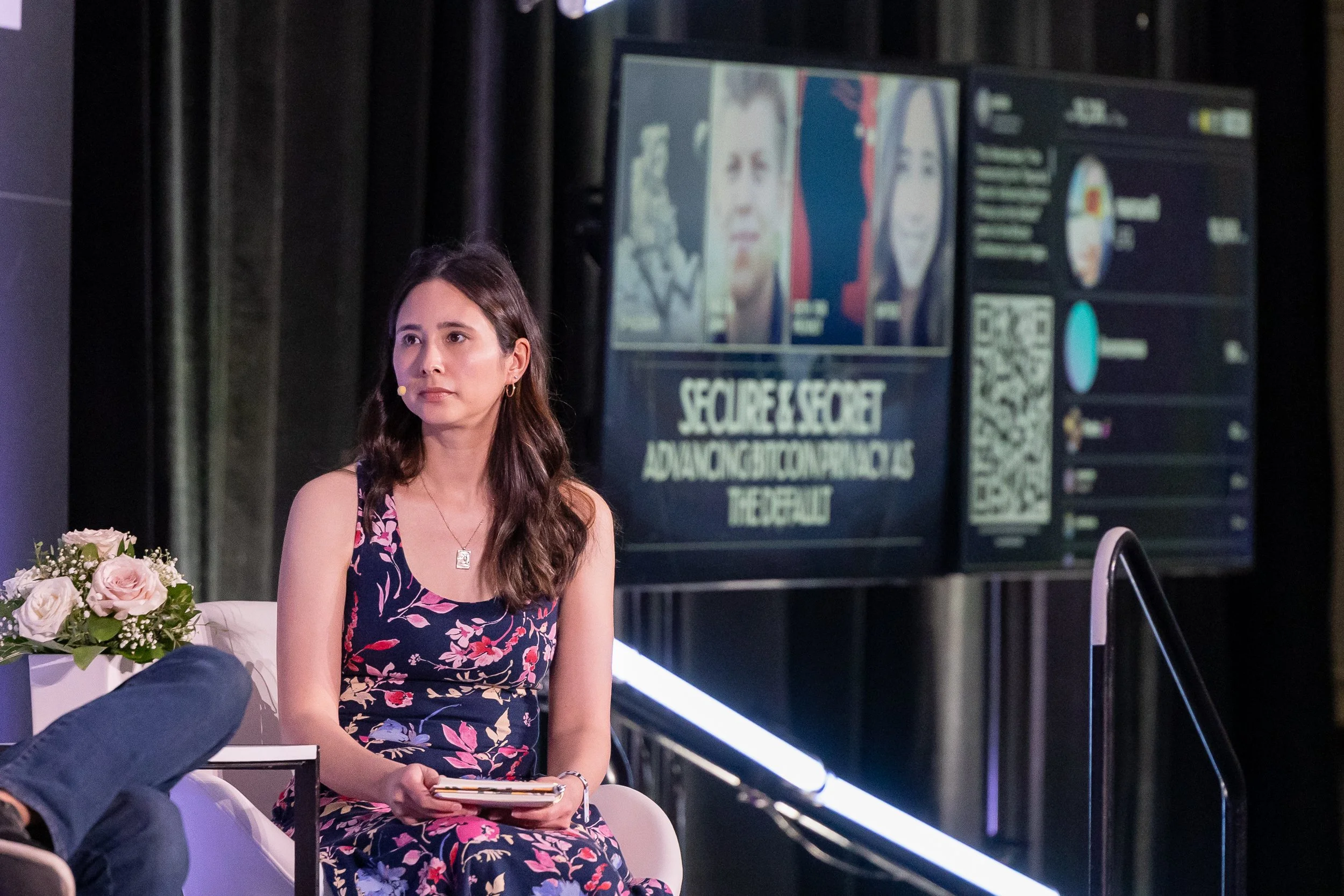 A woman sitting at an event with a flower arrangement in front of her and a screen displaying faces and text behind her.