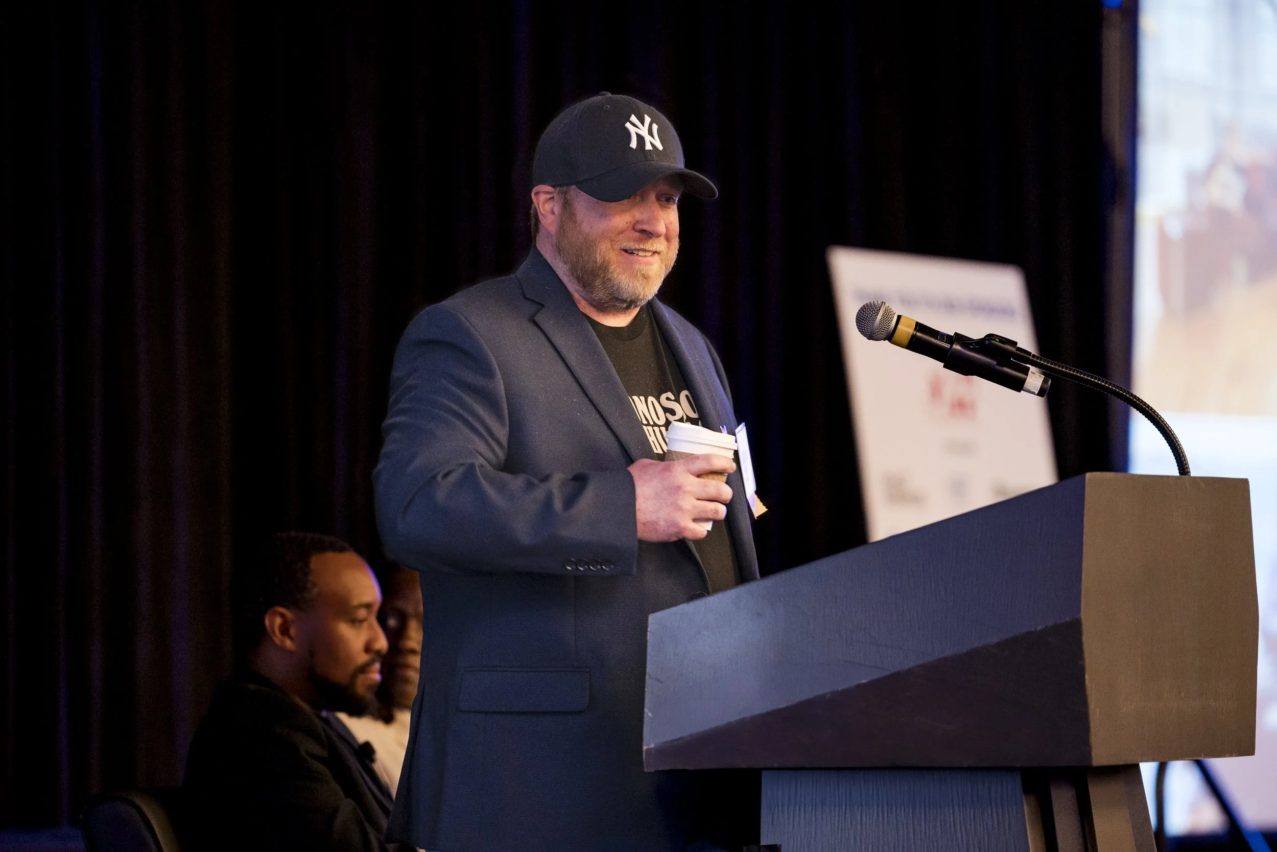 A man in a black New York Yankees cap, dark blue blazer, and T-shirt speaking at a podium with a microphone, holding a disposable coffee cup, in a dimly lit conference room with two seated men in the background.