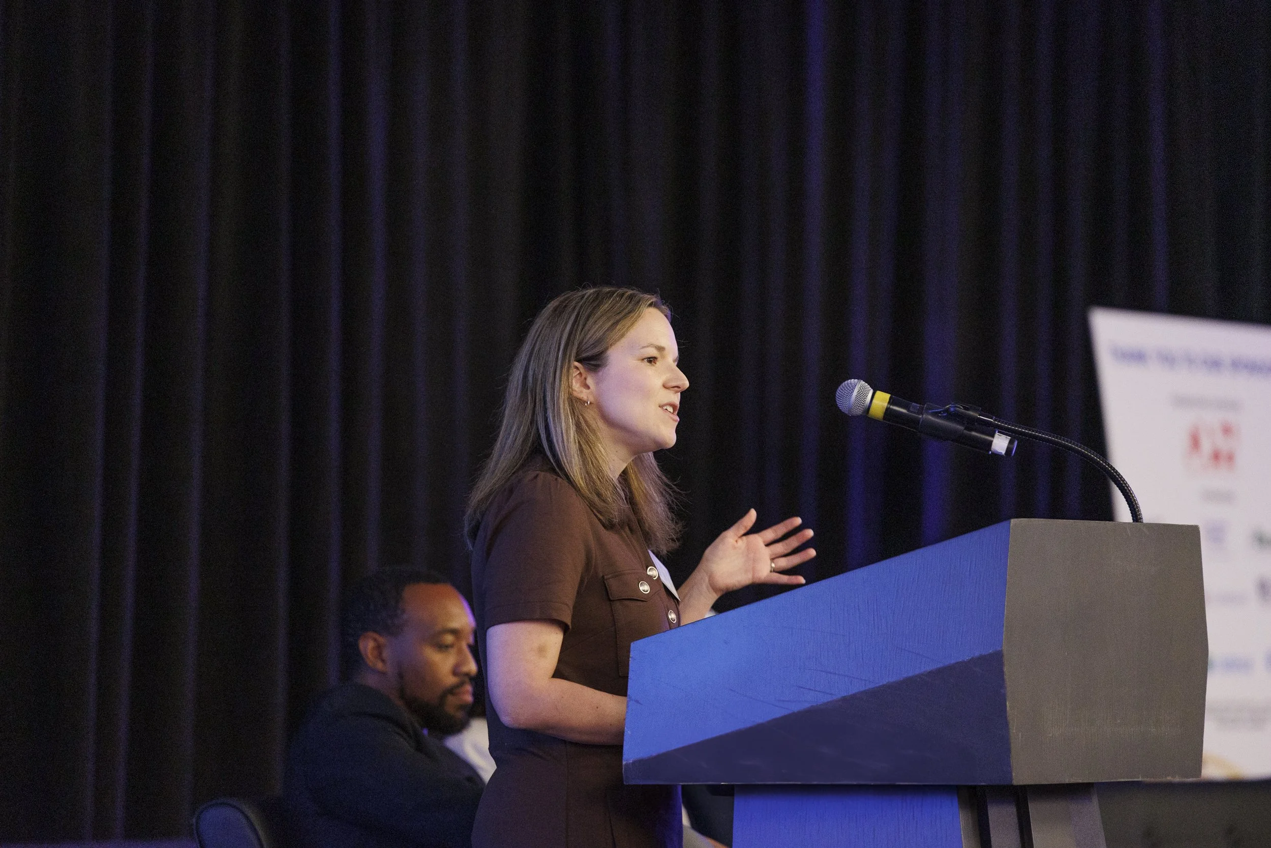 A woman speaking at a podium during a conference or event with a person seated behind her and a banner in the background.