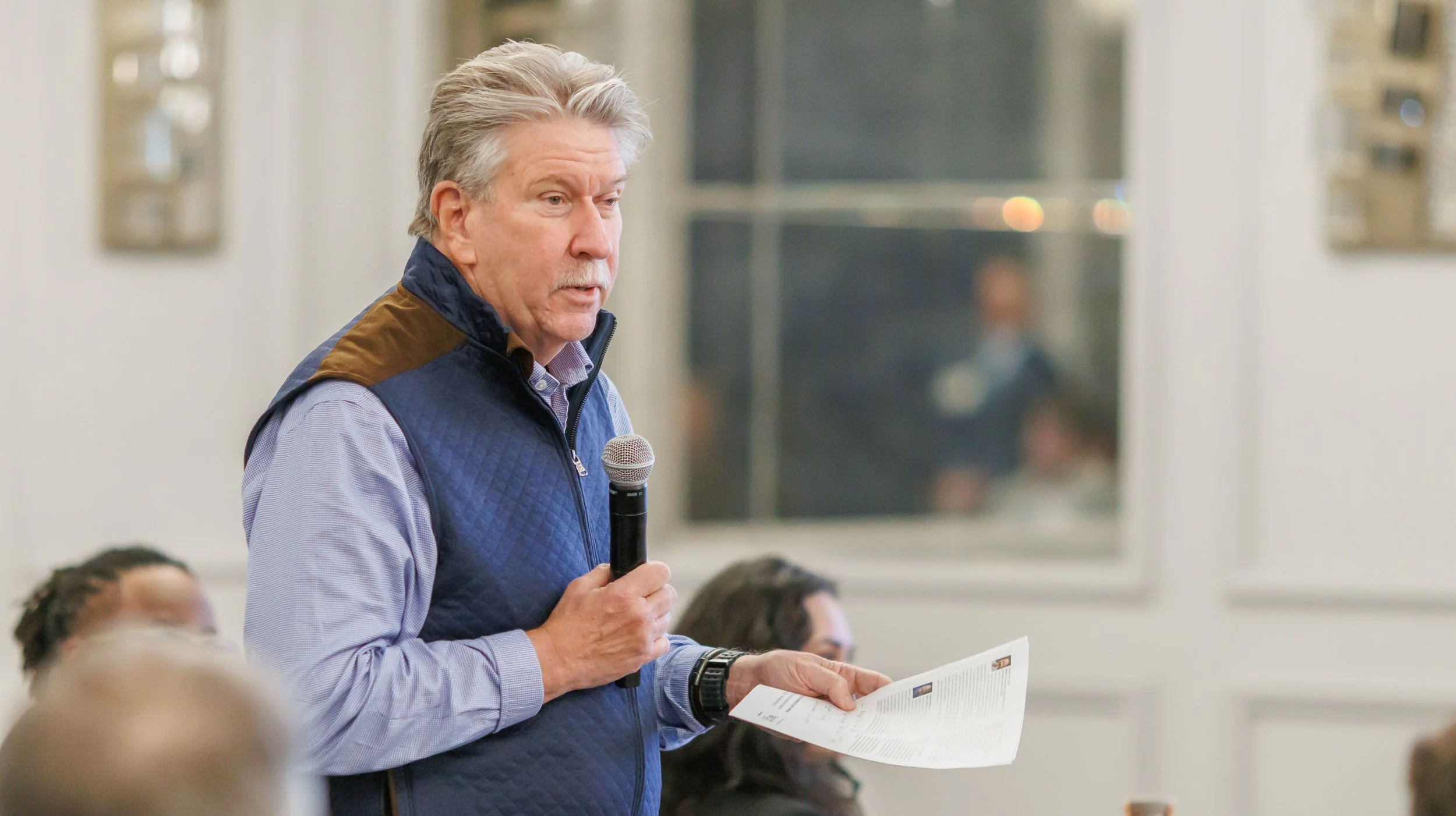 An older man with gray hair and a mustache holding a microphone and a piece of paper, speaking at a gathering in an indoor setting, with a blurred background of people and framed pictures on the wall.