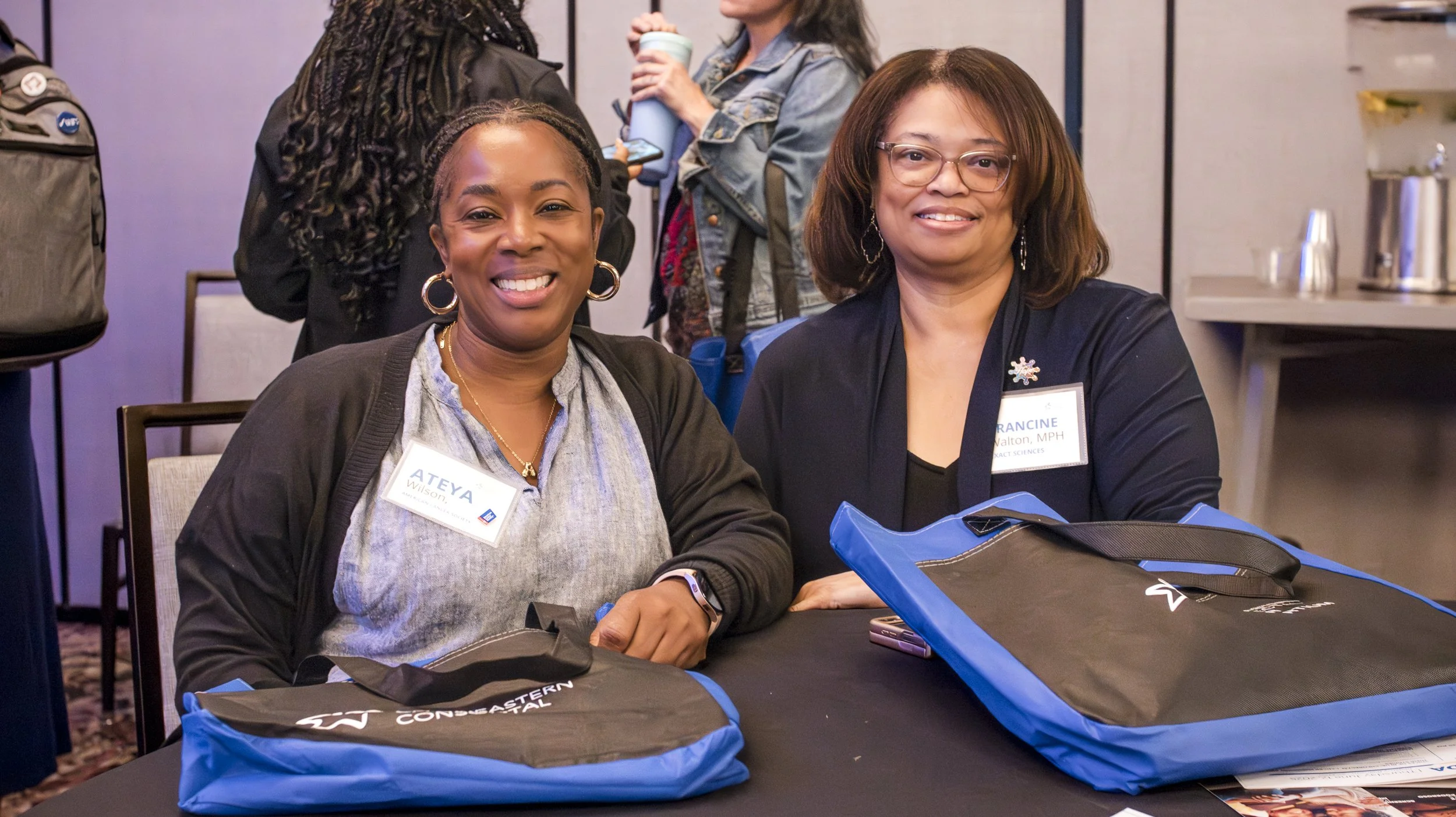 Two women sitting at a table smiling, wearing name tags, with conference bags in front of them, at a professional event or conference.