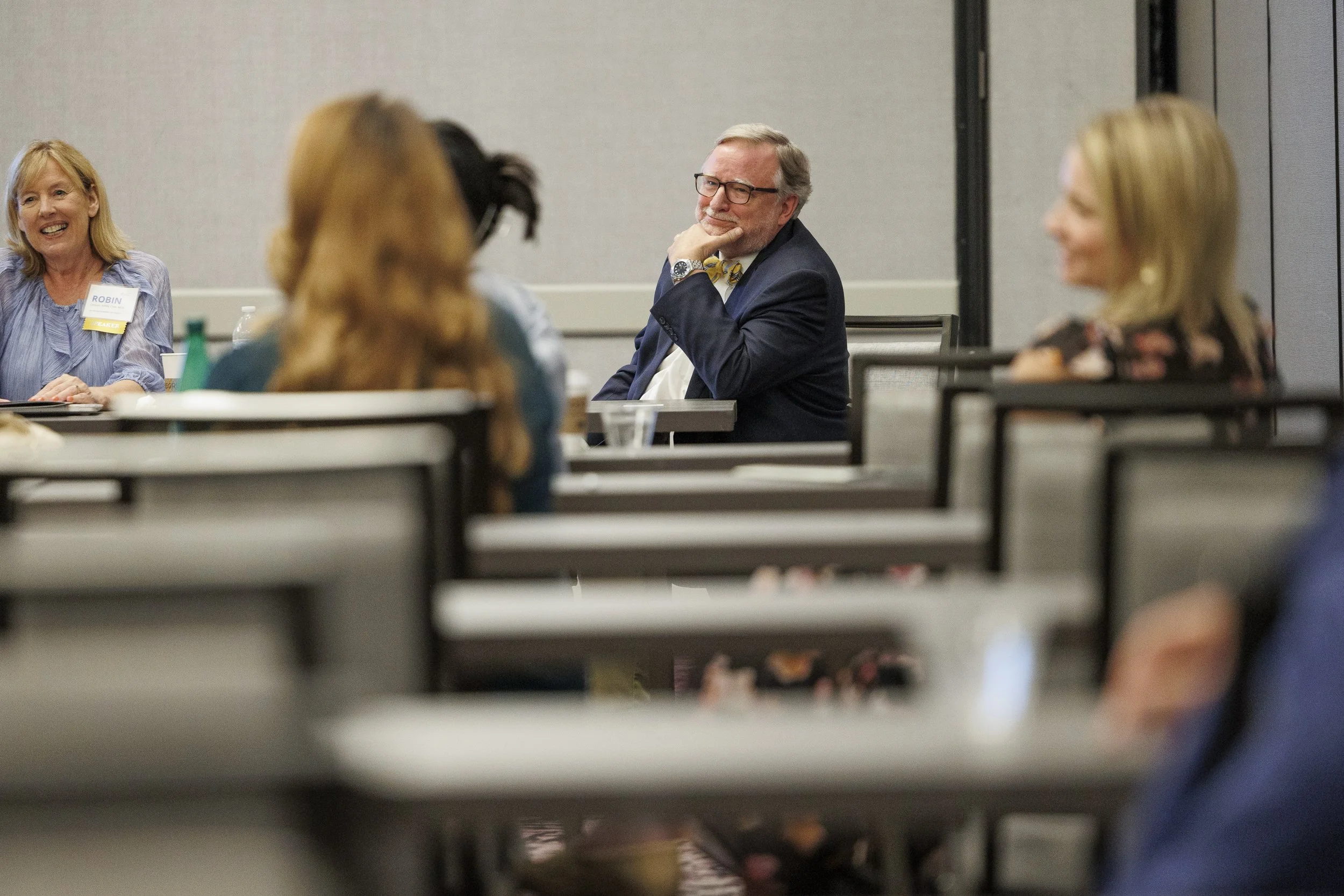 People attending a conference sitting at tables, listening to a speaker or presentation, with one man in glasses and a bowtie smiling thoughtfully.