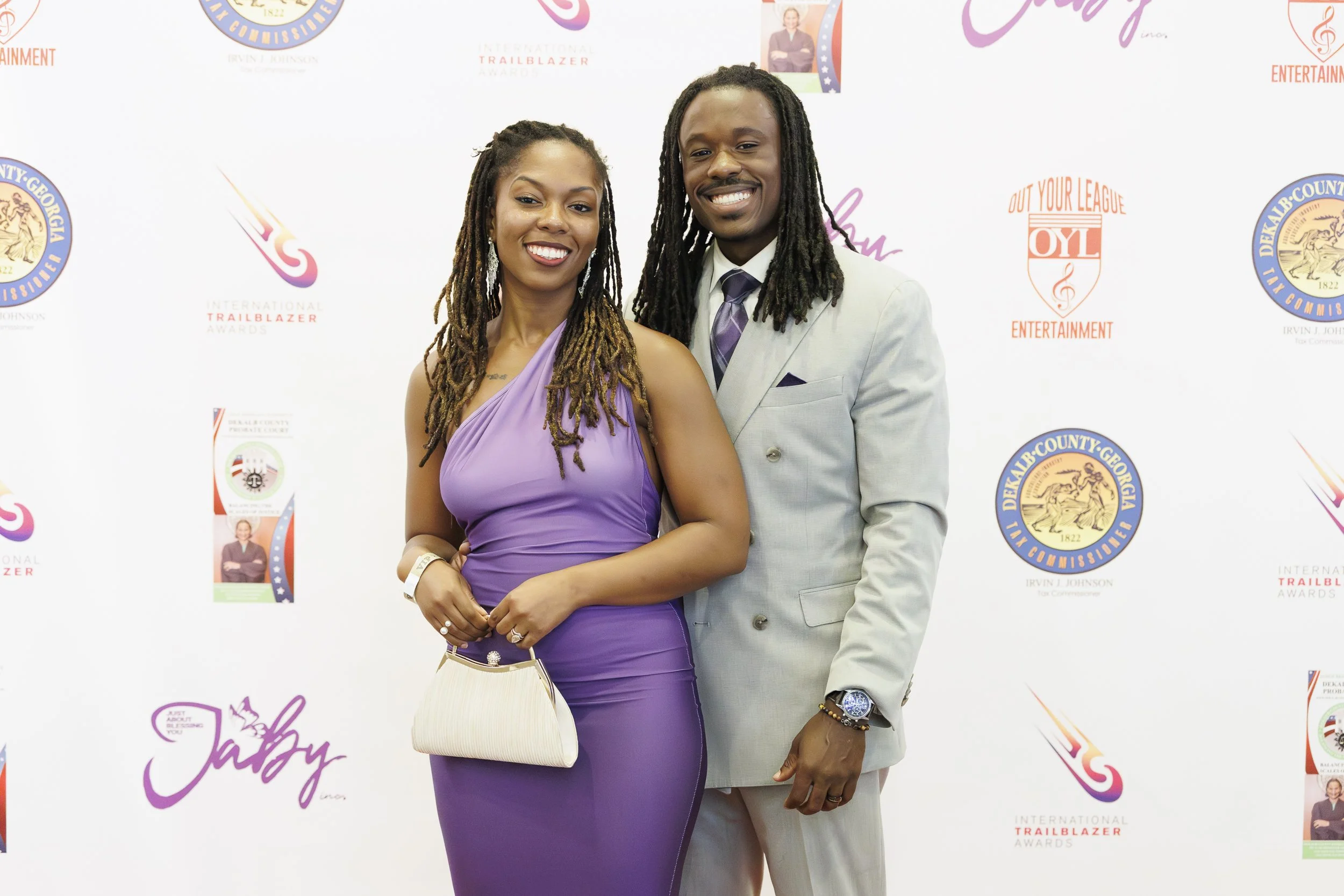 A smiling woman in a purple dress and a man in a light gray suit with a purple tie, standing together at an event with a step-and-repeat backdrop featuring logos and text.