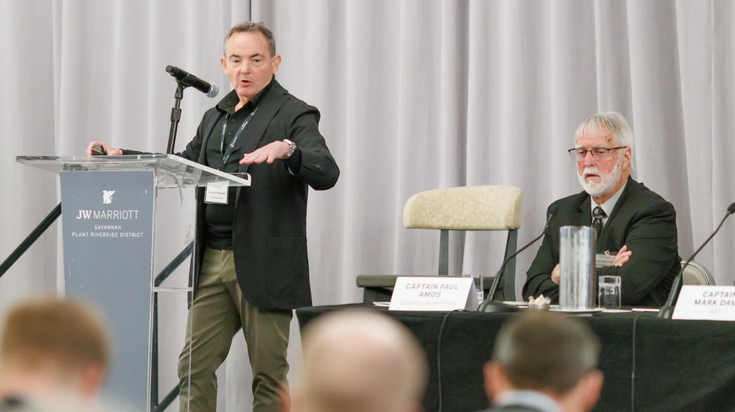 Man in black blazer speaking at a podium during a conference in a hotel conference room, with another man sitting at the table to the right.