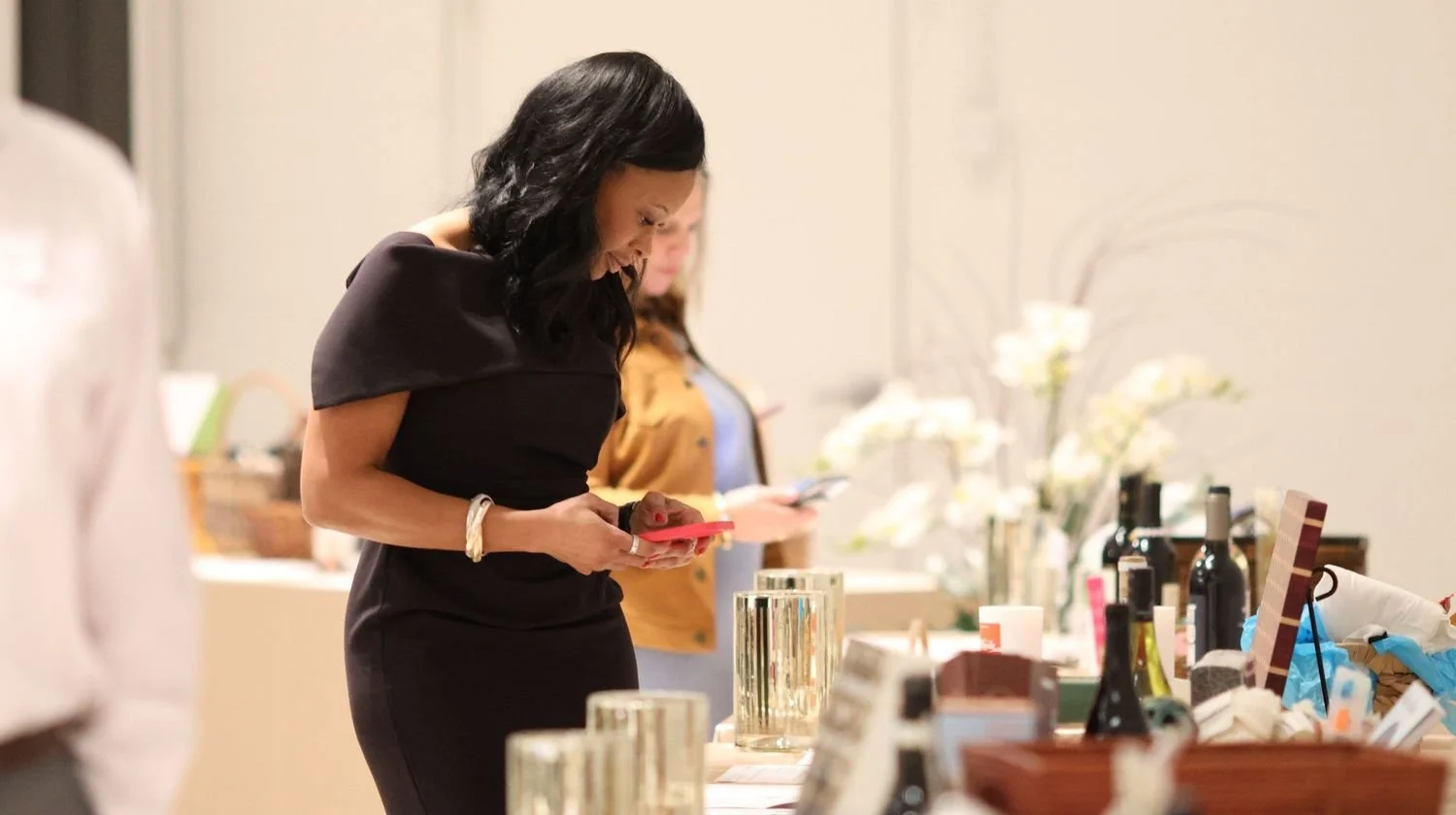 Woman in black dress looking at her phone at a table with various bottles and decorative items.